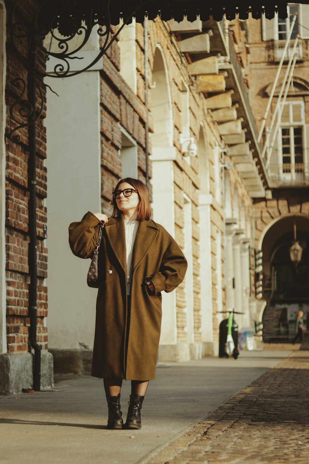 a woman standing on a sidewalk wearing a brown coat