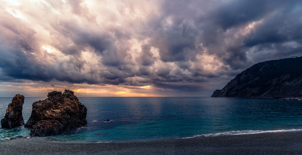 a cloudy sky over the ocean with rocks in the foreground