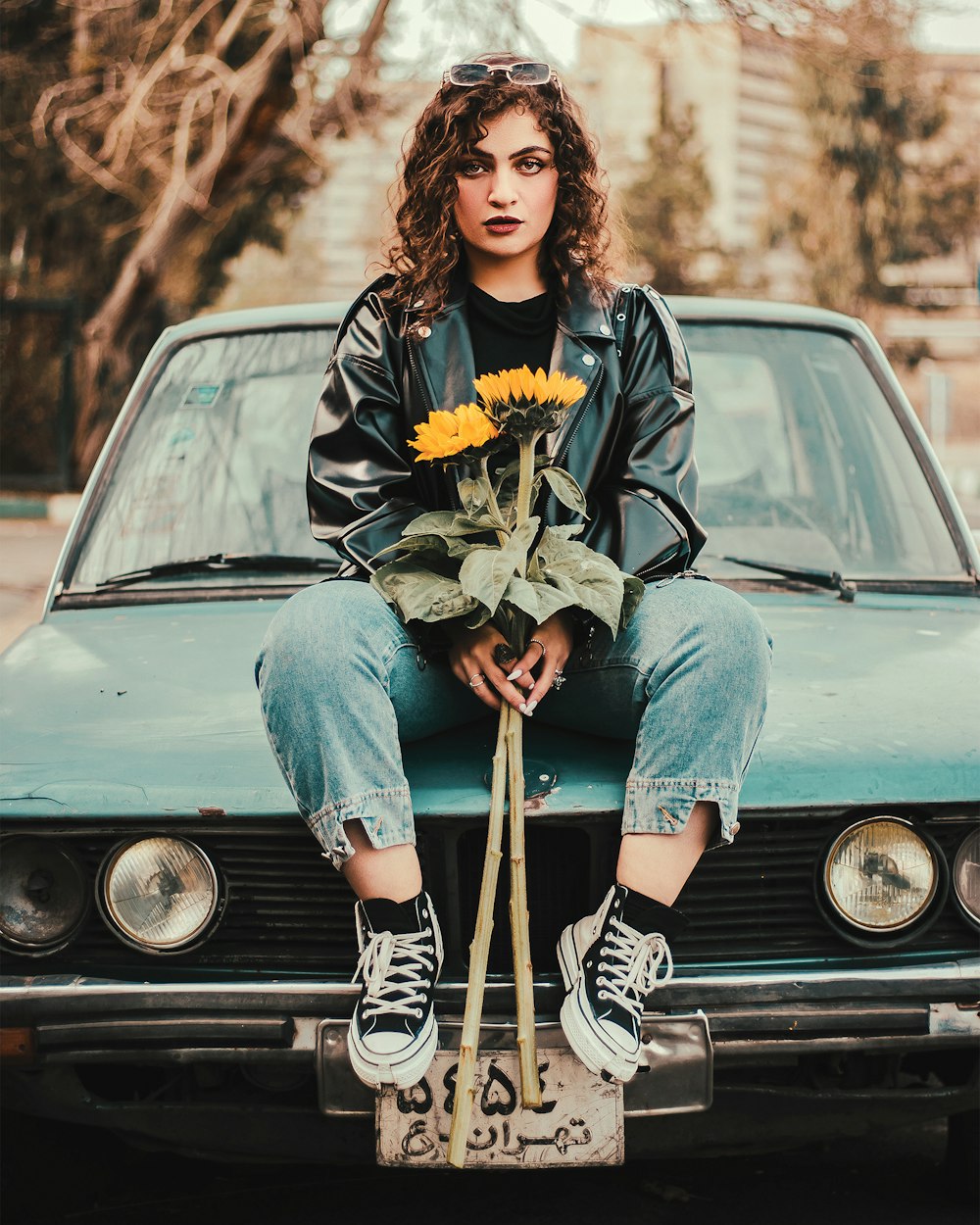 a woman sitting on the hood of a car with a bouquet of sunflowers