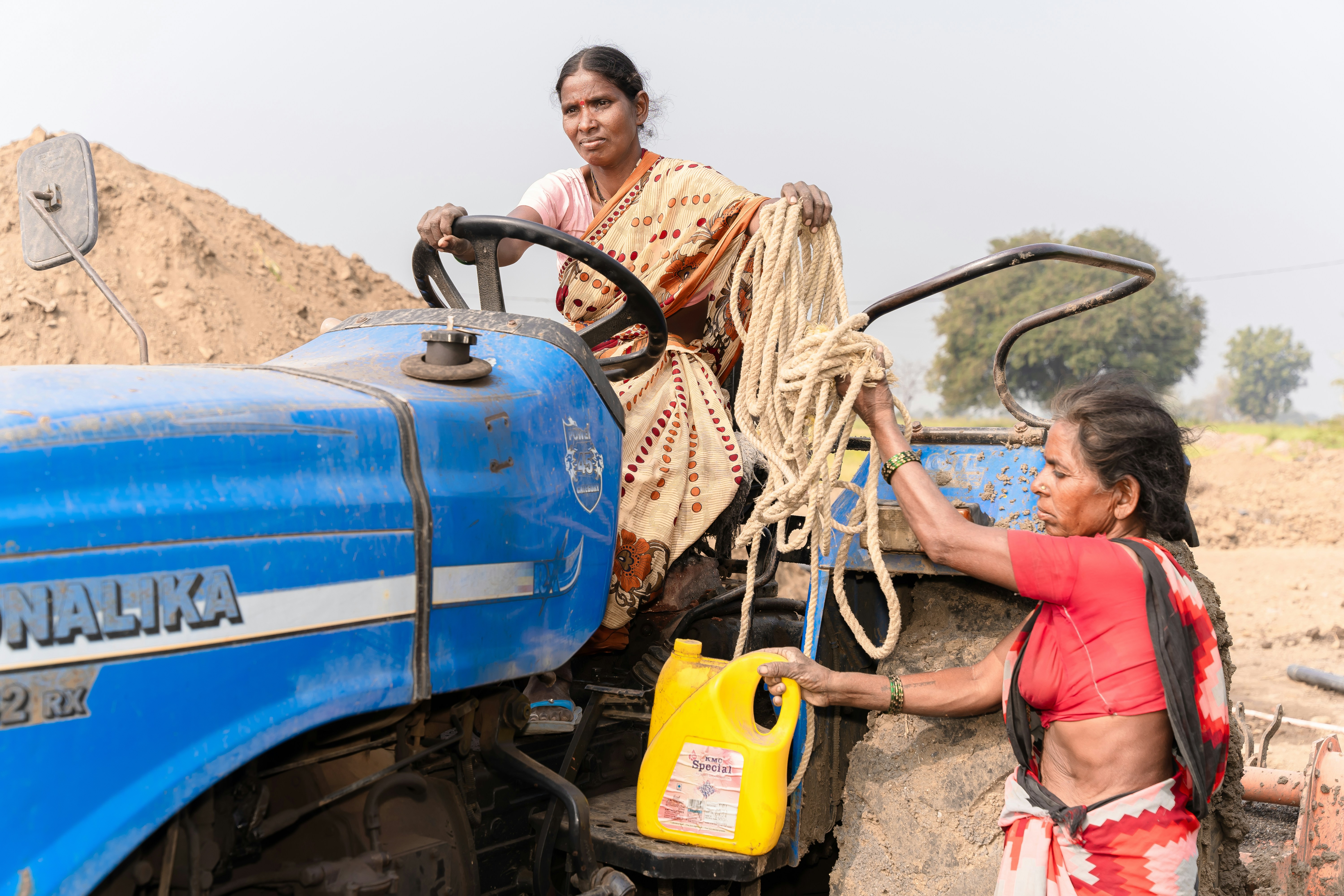 एक भारतीय गाँव में महिलाएं स्वच्छ पेयजल के लिए हैंडपंप का उपयोग कर रही हैं, पृष्ठभूमि में हरे-भरे खेत दिख रहे हैं।