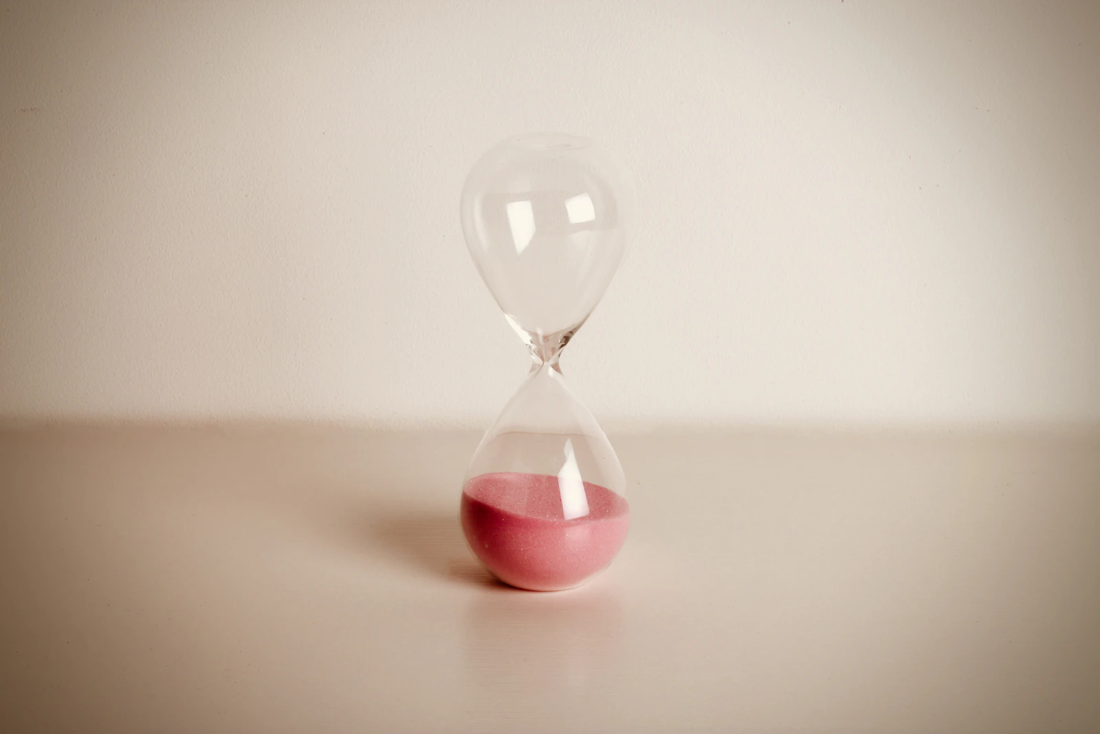 A small hourglass with rose-colored sand on a warm cream surface