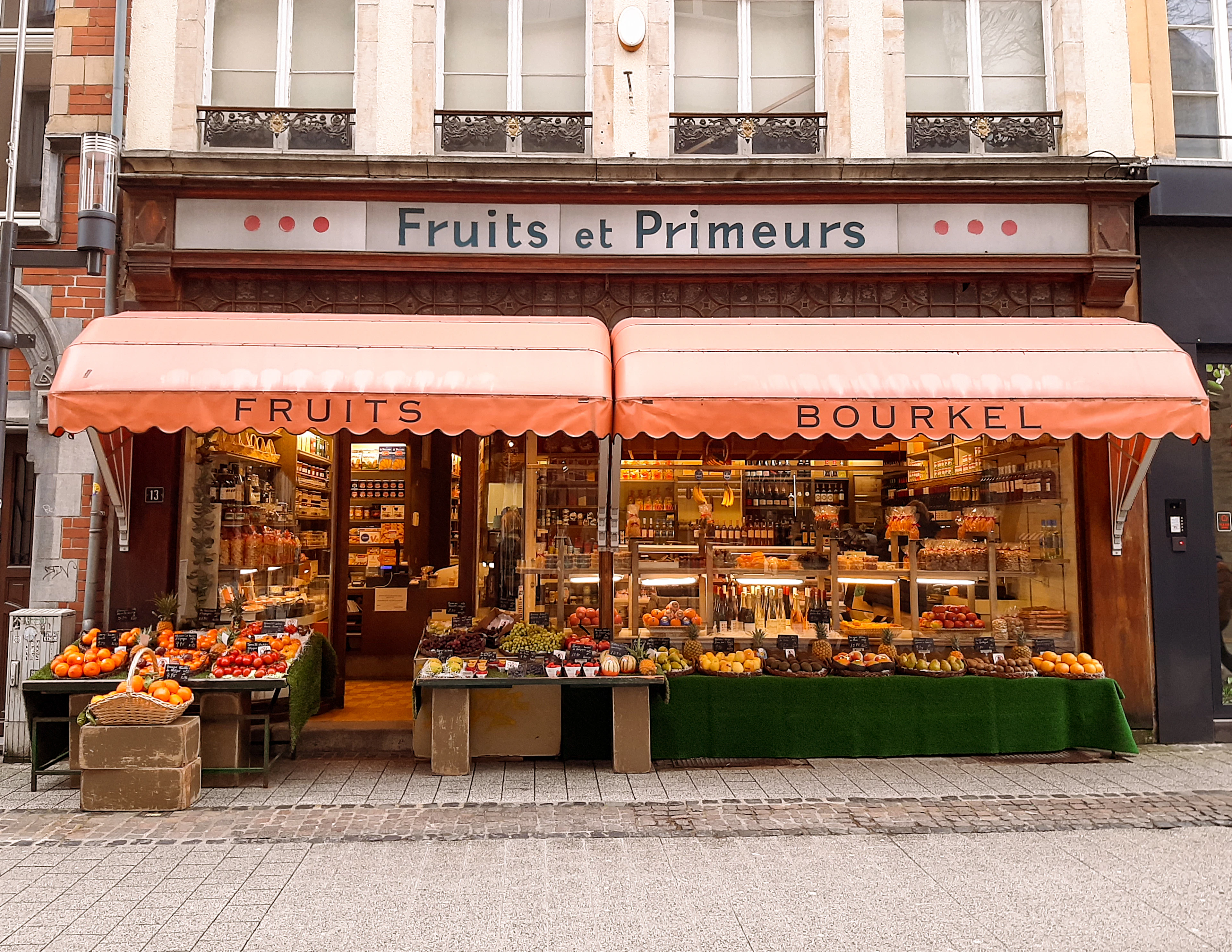 A greengrocer's shopfront under orange awnings lettered 'Fruits et Primeurs', with open crates of oranges, apples, peppers and greens stacked on trestle tables across the pavement and jars and produce filling the windows behind.