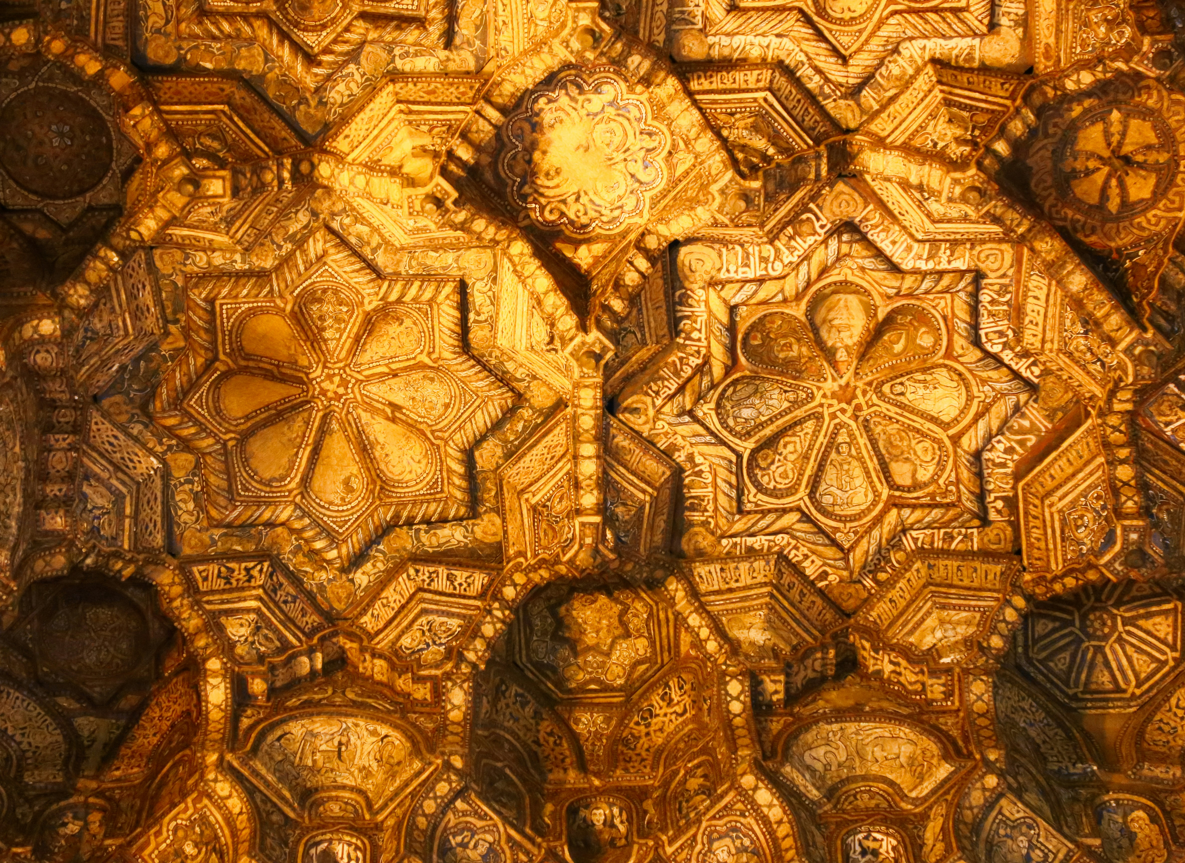 This image features a close-up of an ornate, gilded ceiling with complex geometric patterns and arabesque designs, reflecting a high level of craftsmanship and historic architectural beauty.