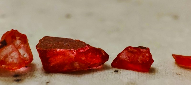 A row of rough, translucent red stones lit from behind on a pale cloth, the largest one flat-topped with a dusty surface