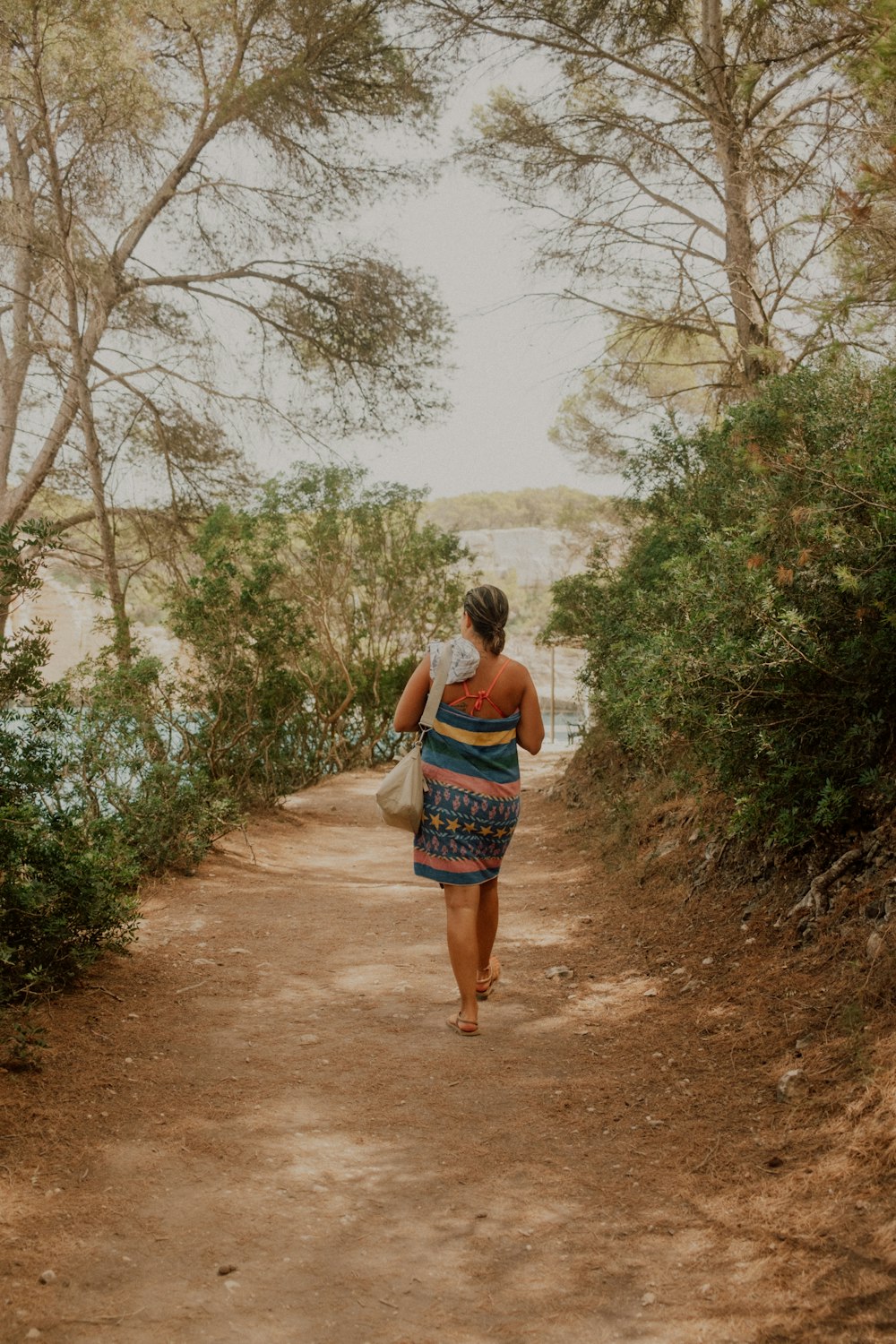 a woman walking down a dirt road next to a forest