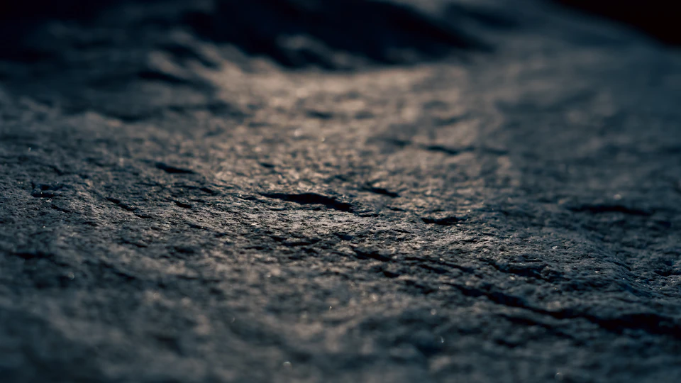 The image shows a close-up view of a rock surface, with a dark gray hue dominating the scene. The rock's texture is rough and uneven, suggesting it has been worn down over time by natural elements such as wind and water. The rock appears to be in good condition overall, but there are some small chips missing from its surface, indicating that it has experienced some wear and tear.