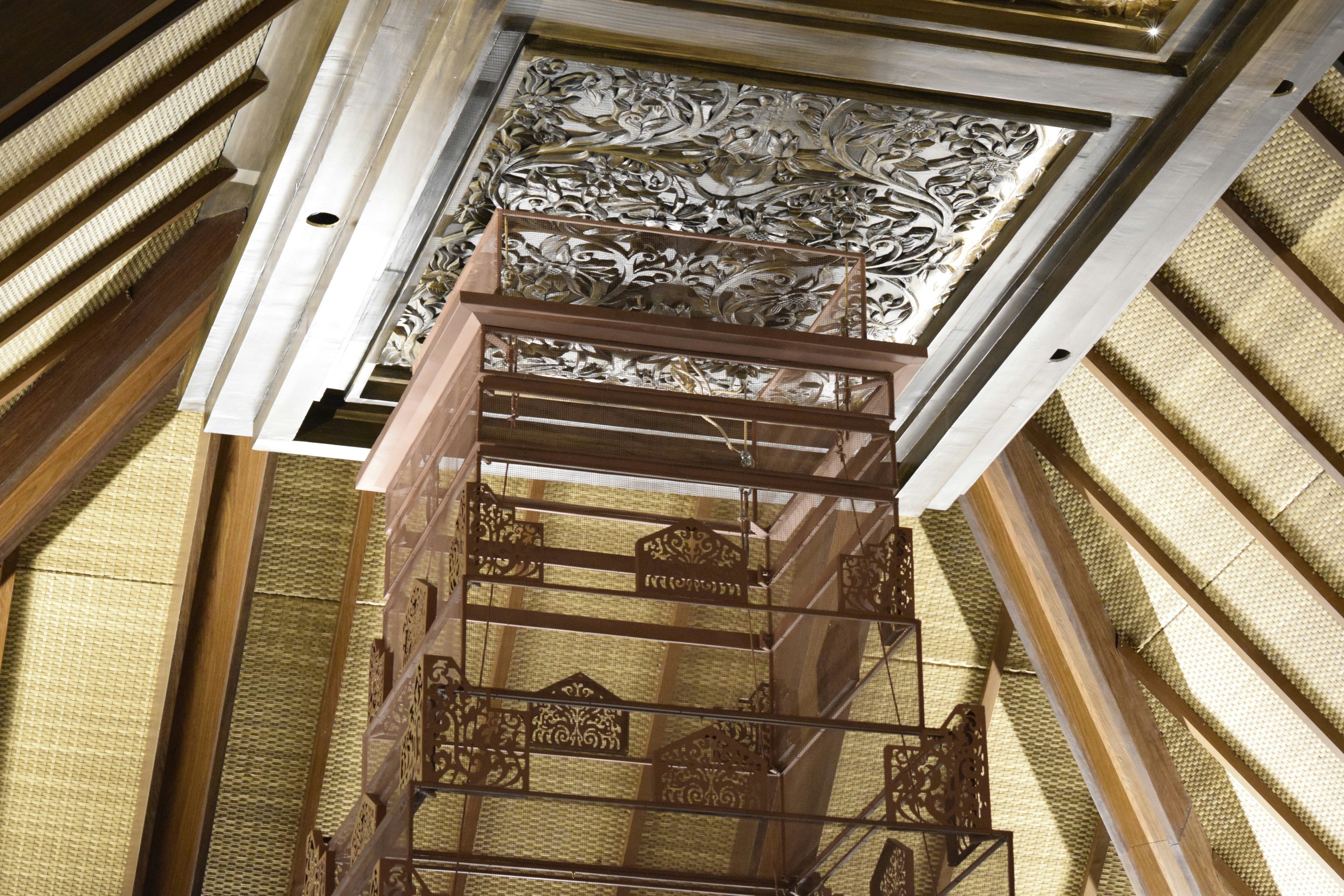 Ornate traditional interior features a finely carved wooden ceiling, a multi-tiered bronze chandelier-like structure, and textured woven walls under an elaborate beamed roof.