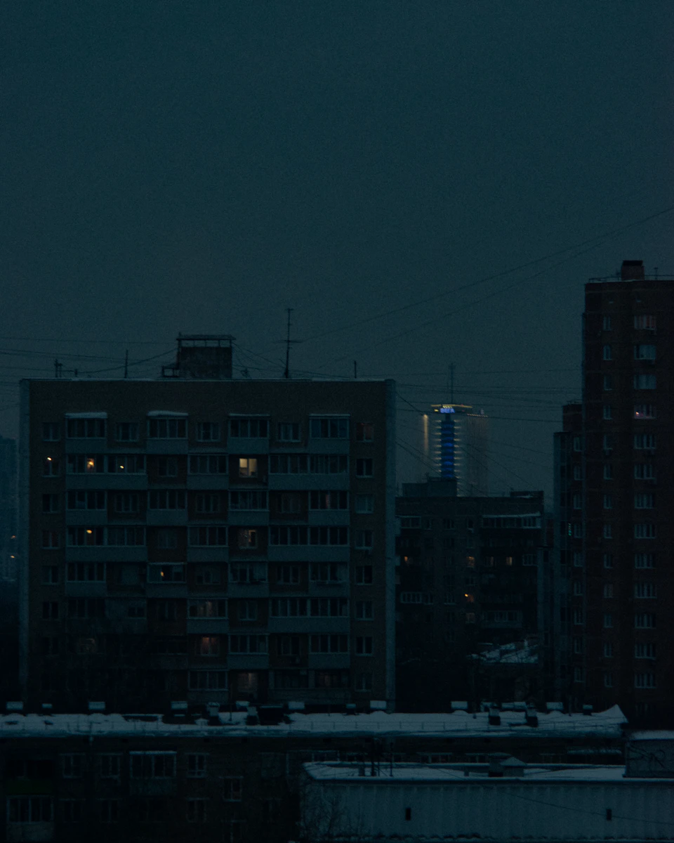 The image depicts a cityscape at night, with tall buildings illuminated against the dark sky. The tallest building is a modern skyscraper with a blue glass facade that reflects the surrounding lights. Below it, there are several other buildings, including an older brick structure and another modern high-rise. The streetlights cast long shadows on the snow-covered ground below, creating a contrast between the illuminated structures and the dark sky.