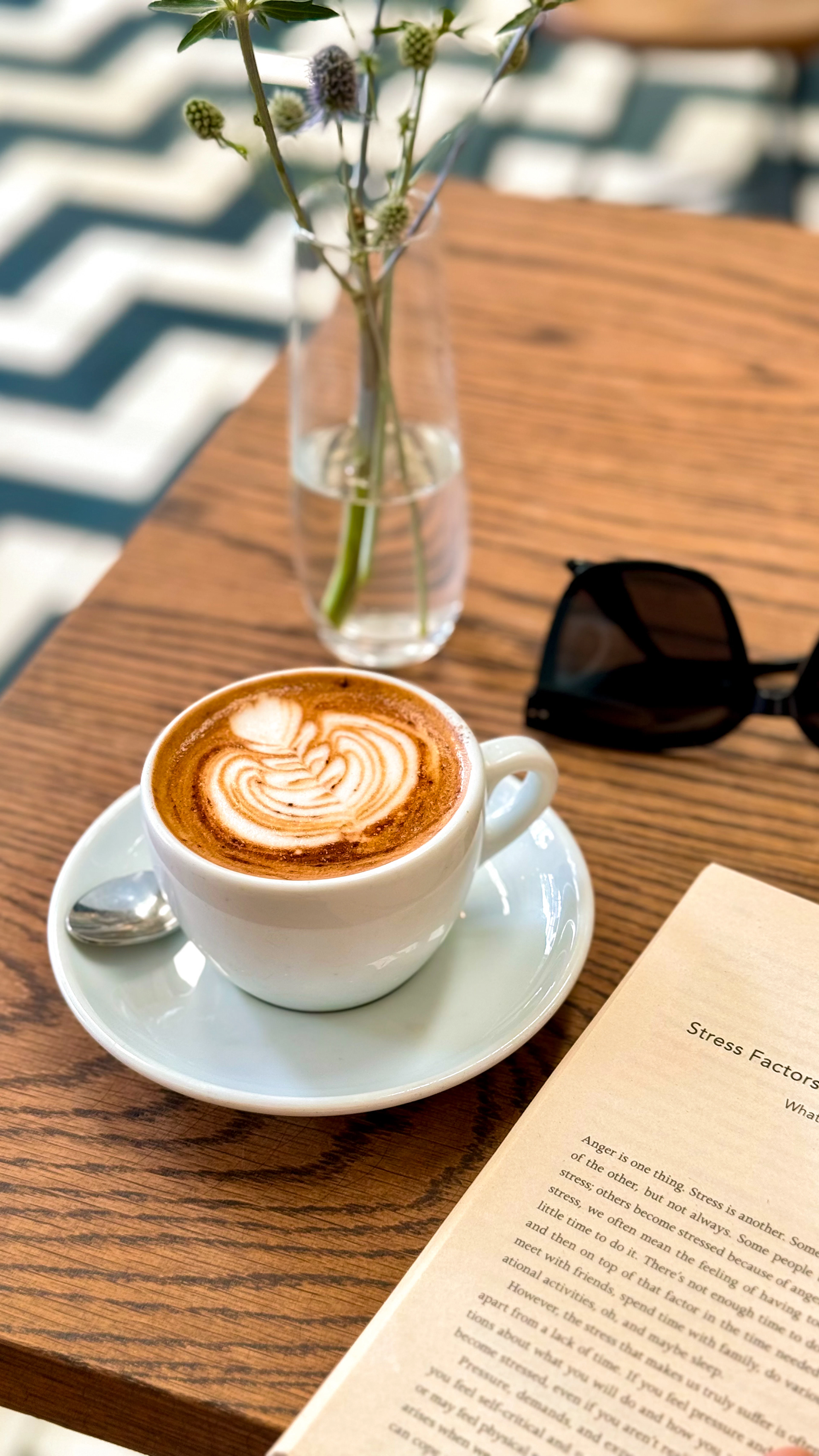 A cappuccino with rosetta latte art on a saucer, next to sunglasses and an open paperback on a sunlit timber café table