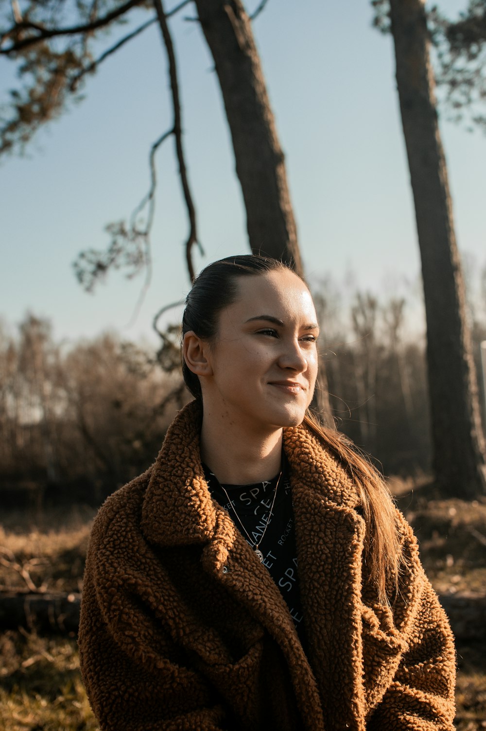 a woman in a brown coat standing in front of some trees