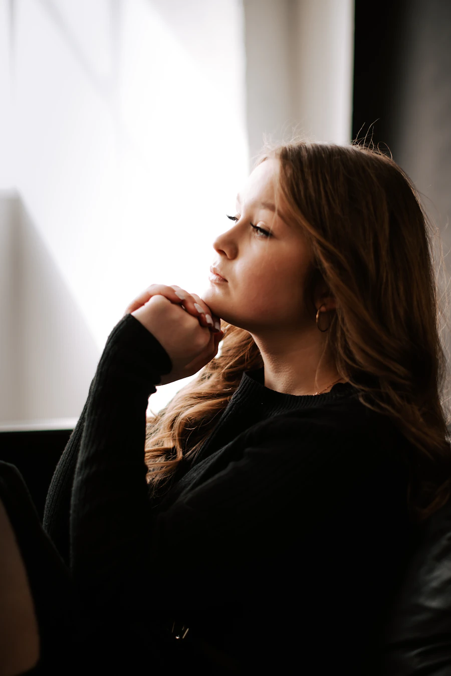 An adult sits in profile by a softly lit window, hands gently clasped beneath the chin in a settled moment of focus