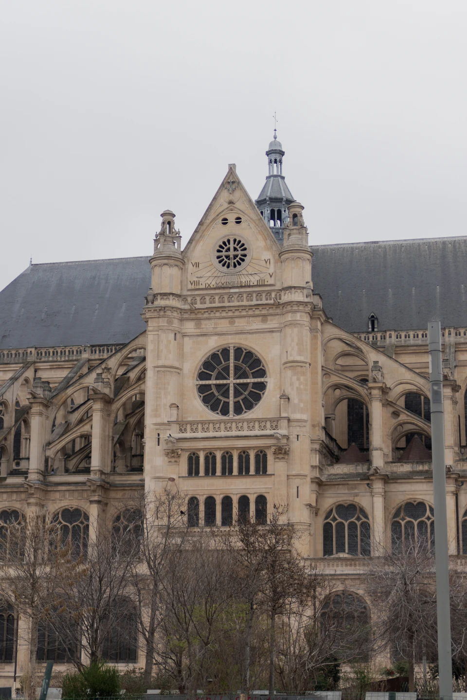 The image shows a large, ornate stone building with a clock tower and a bell tower on its roof. The clock face is white, contrasting against the gray sky above. The building features multiple arched windows that add to its architectural charm. In front of the building, there are trees visible, providing some greenery to the scene.