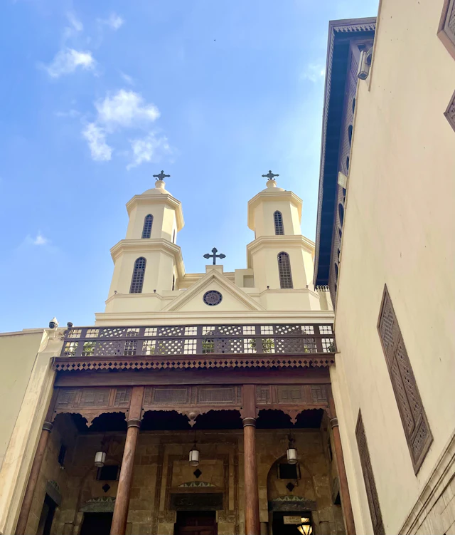 The Hanging Church's cream twin bell towers and rose window seen from below, with its wooden latticed balcony and columns.