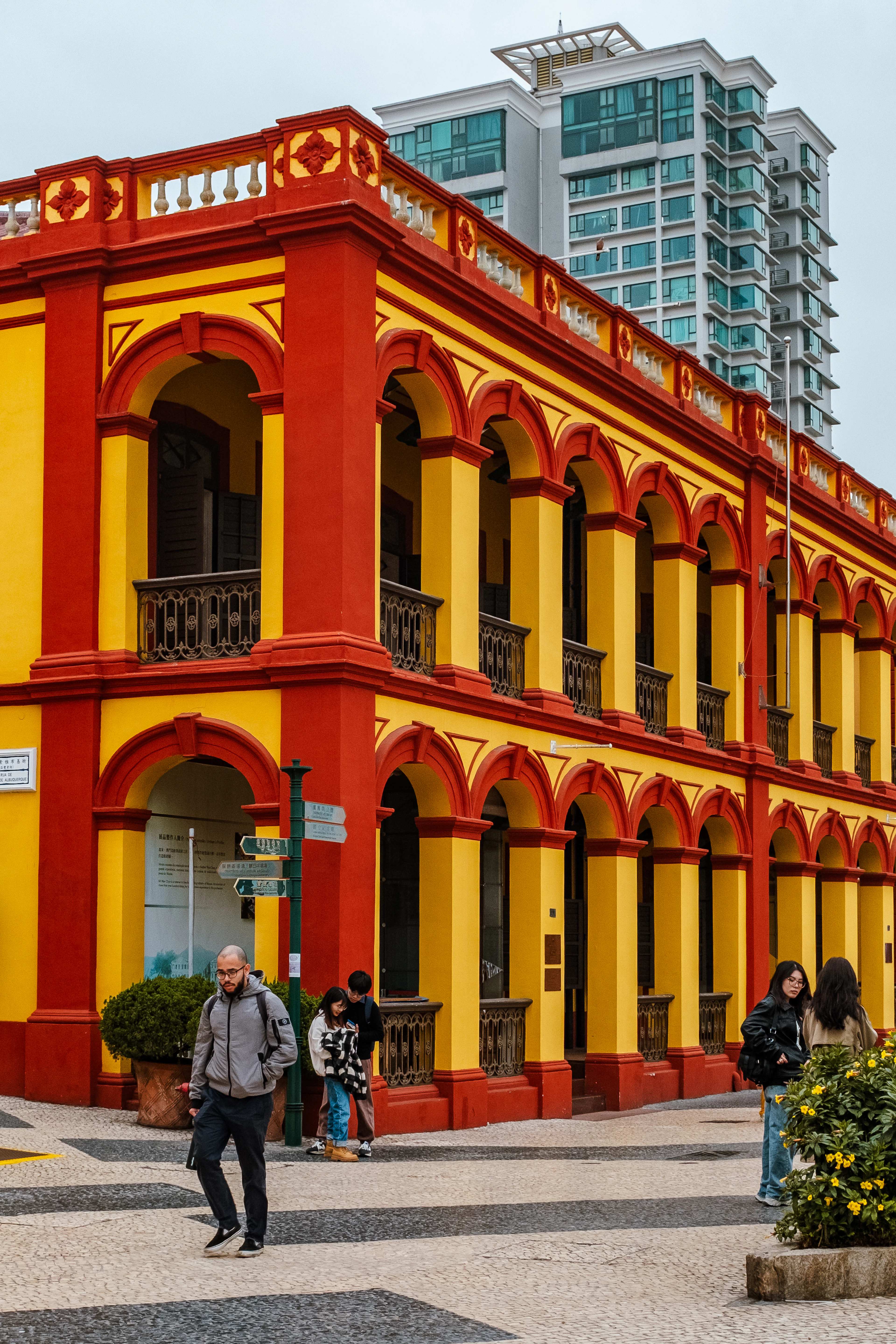A two-story ochre-yellow colonial building with red-trimmed arcades and balconies, pedestrians passing below.