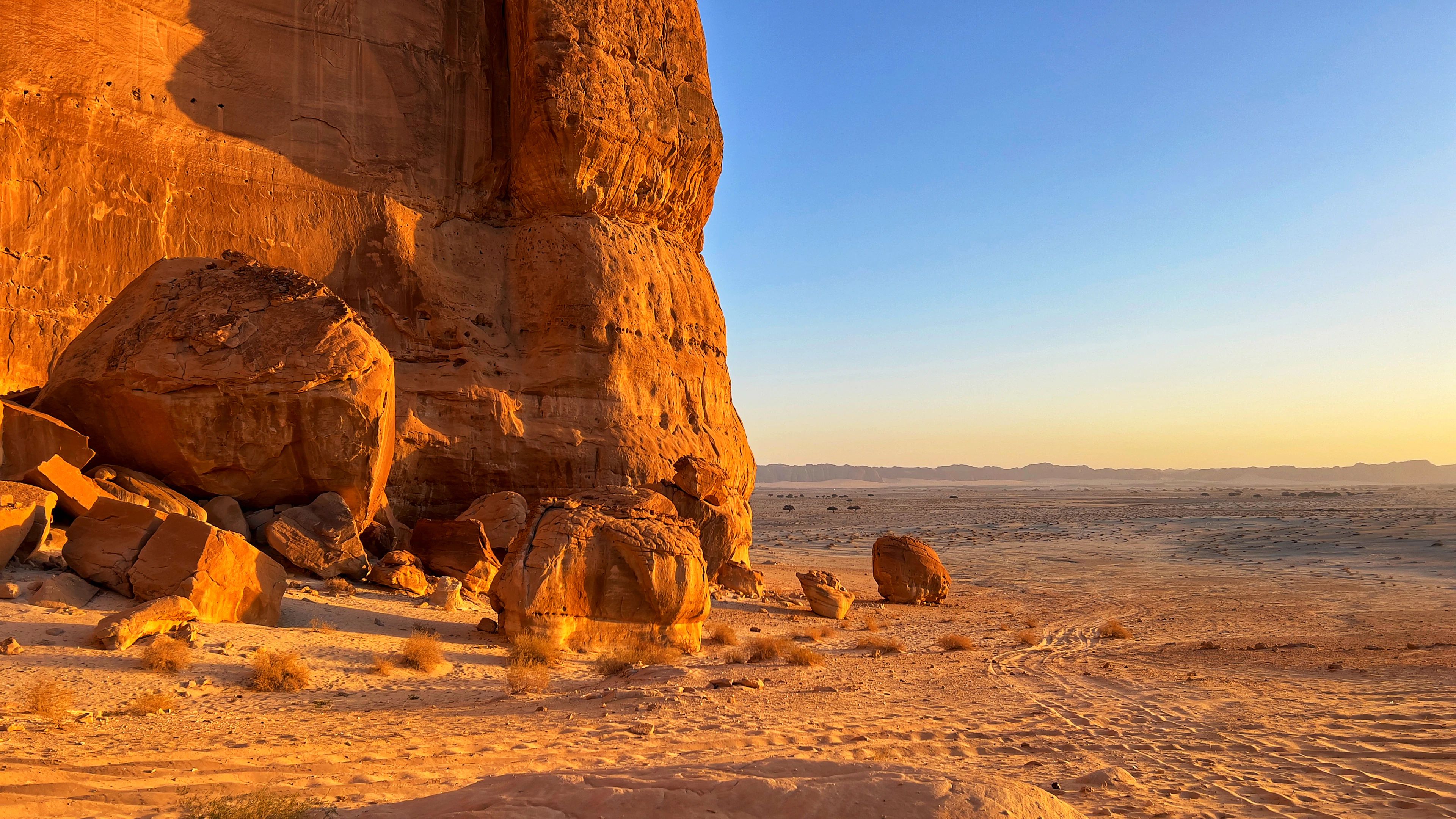 A massive sandstone cliff glows orange at golden hour, its base scattered with boulders and desert scrub stretching to the horizon.