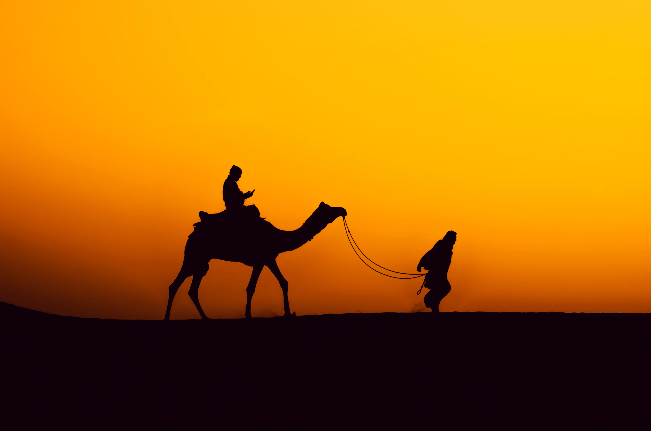 A camel and its handler silhouetted against an orange sunset on the Jaisalmer dunes