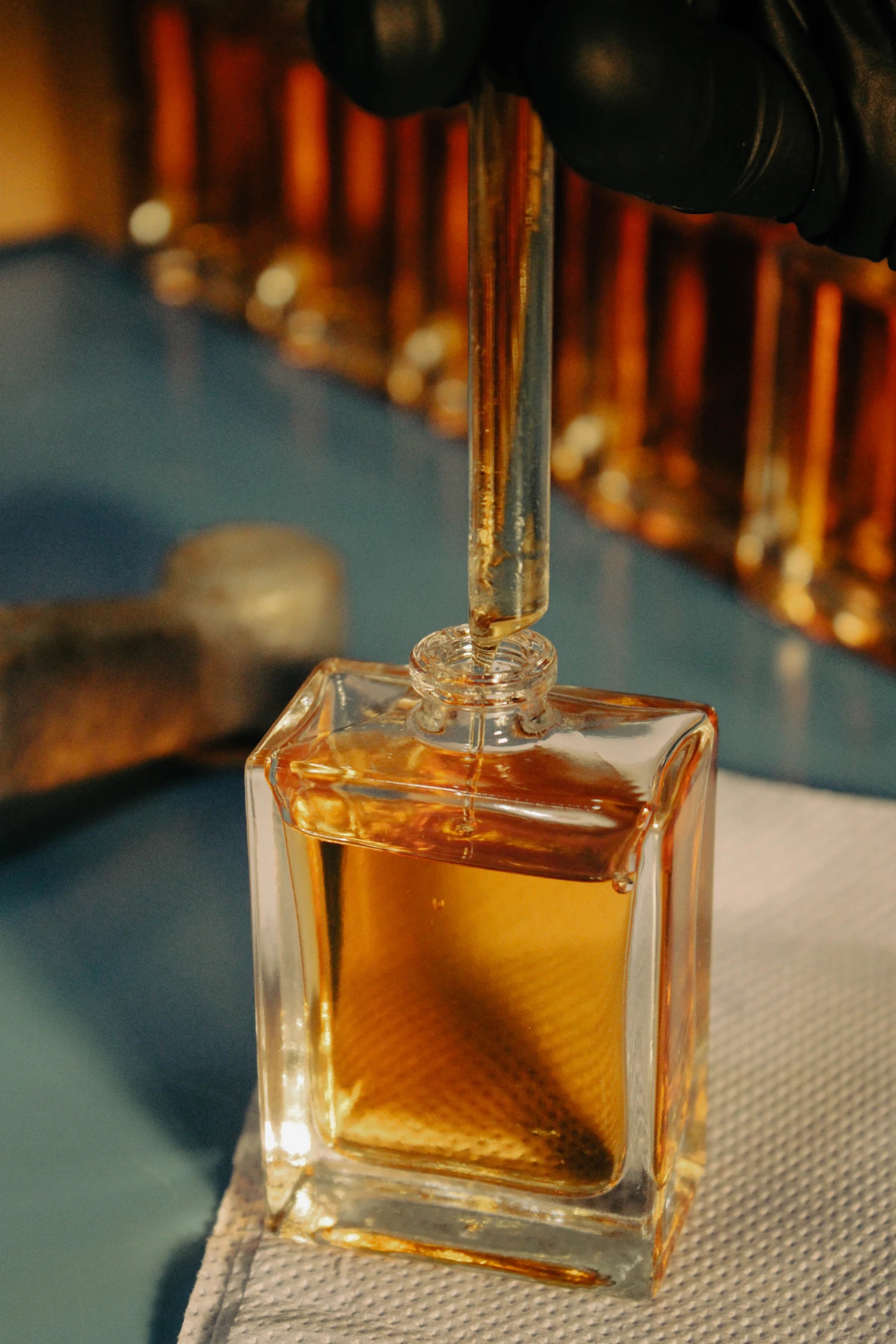 A gloved hand dosing honey-coloured Miele Scuro into a square flacon, a row of amber bottles glowing behind