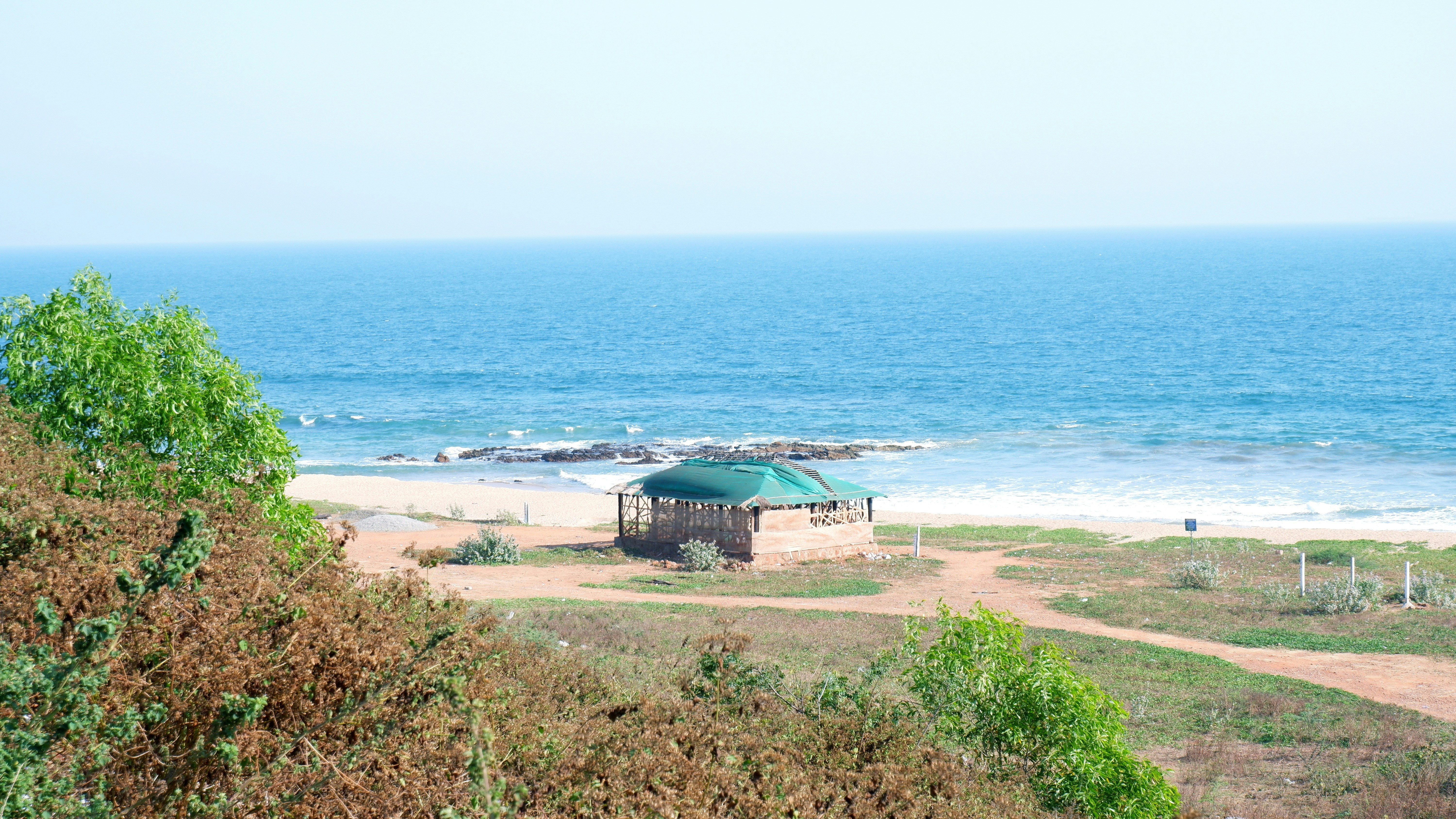 a gazebo sitting on top of a sandy beach next to the ocean, Rushikonda beach is widely known for its Golden Sands, Tidy waves of Bay of Bengal & haven for Sea Surfers. Surrounded by green plants, Rushikonda beach looks picture perfect attracting Tourists & Travellers in huge numbers. With its vast stretch of waters, Rushikonda beach is the perfect place to enjoy water sports like swimming, water skiing and wind surfing. This beach is surrounded by nature lovers and adventure enthusiasts enjoying some beautiful moments amidst the scenic view of the beach with cool breeze caressing them.