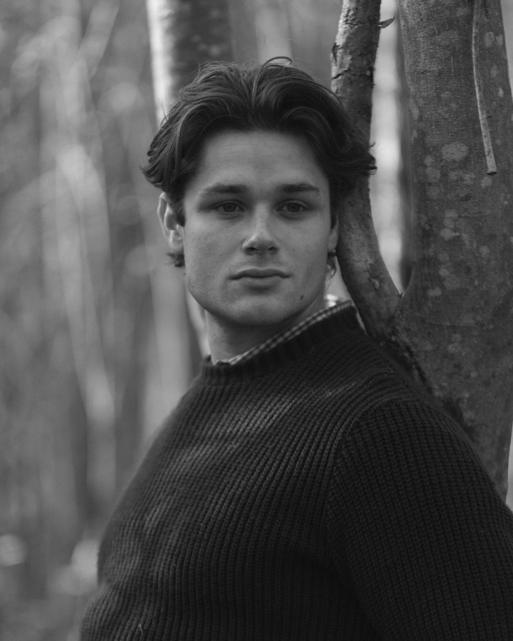 a black and white photo of a young man leaning against a tree