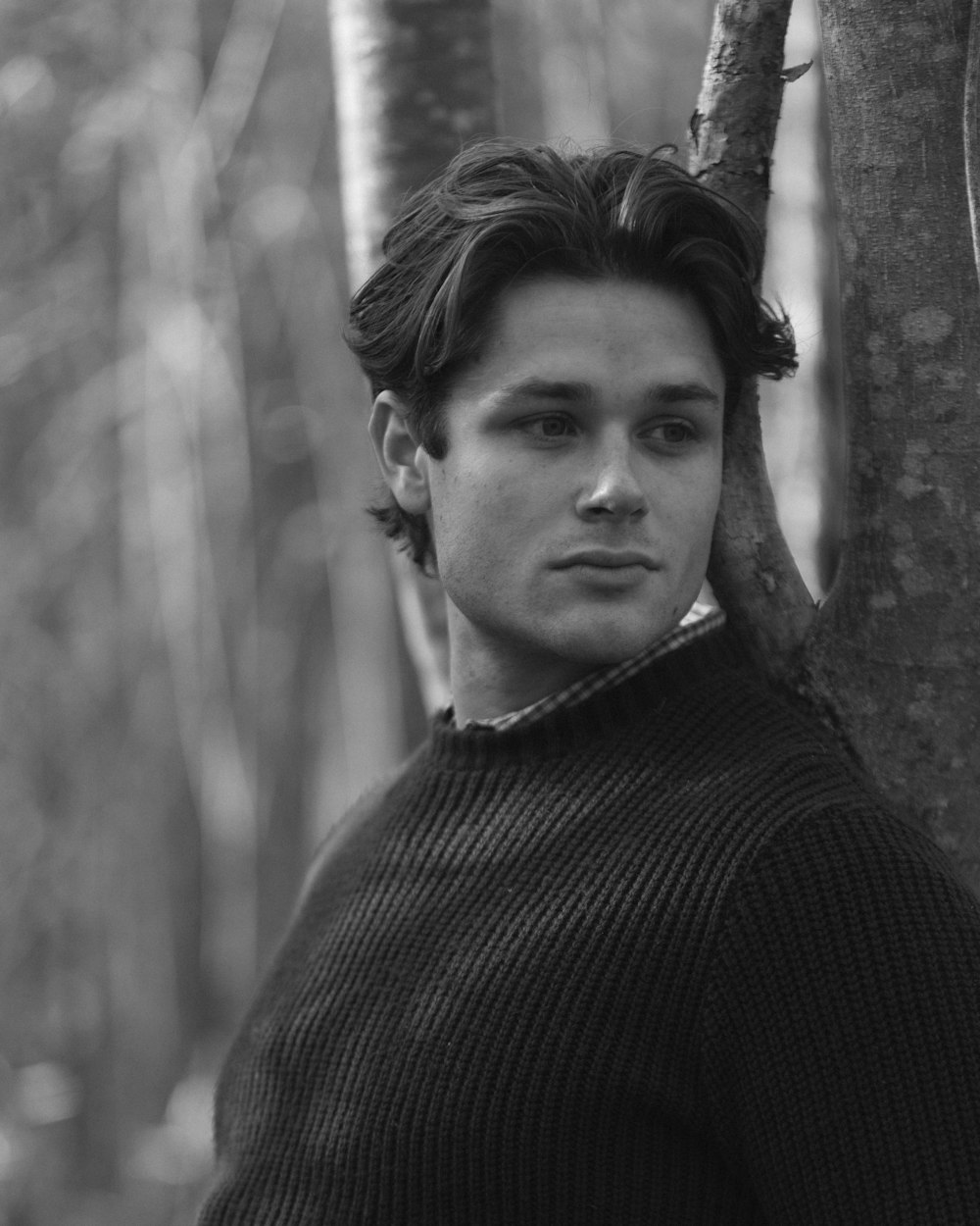 a black and white photo of a young man leaning against a tree