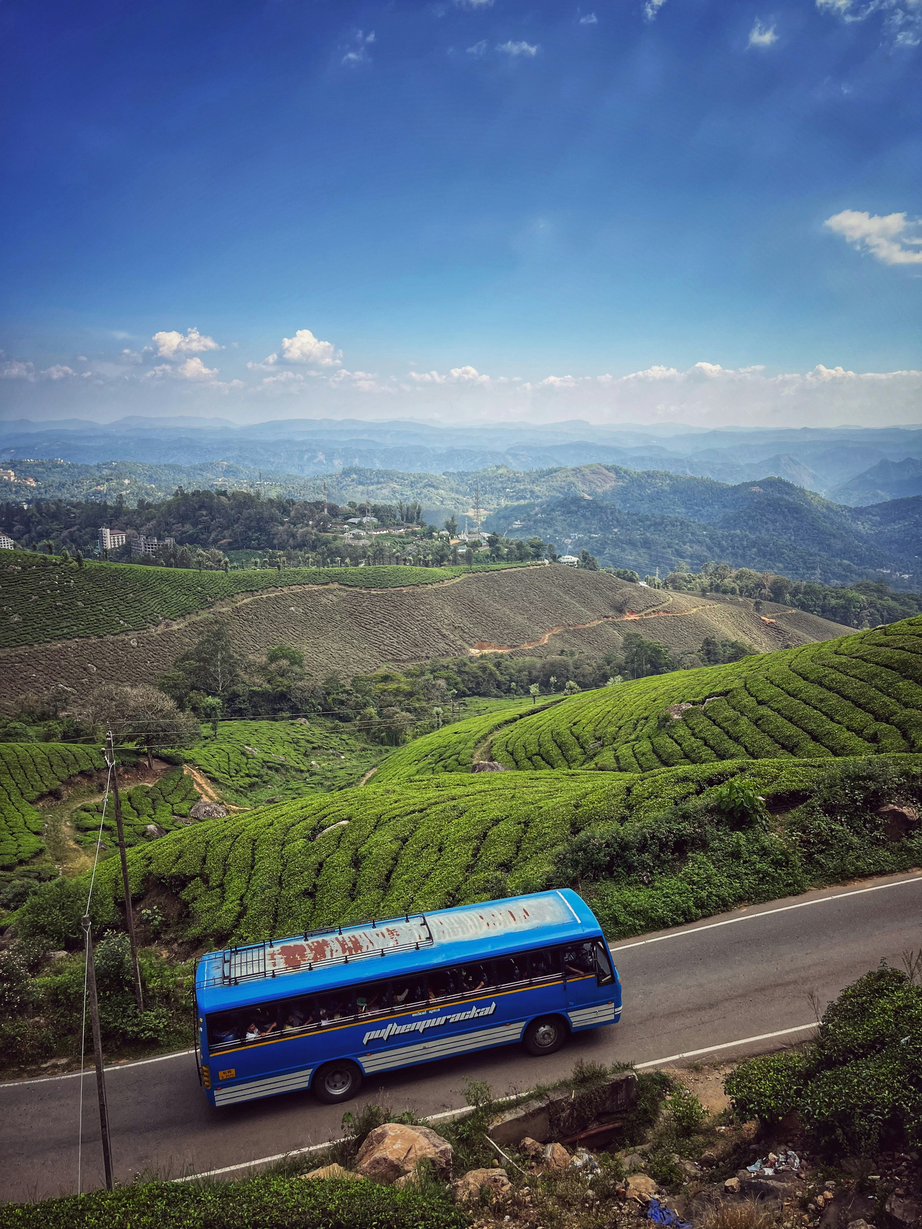 a blue bus driving down a road next to a lush green hillside