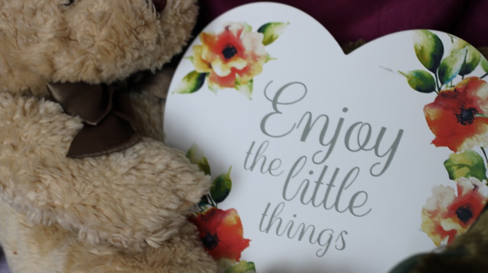 a teddy bear holding a sign that says enjoy the little things