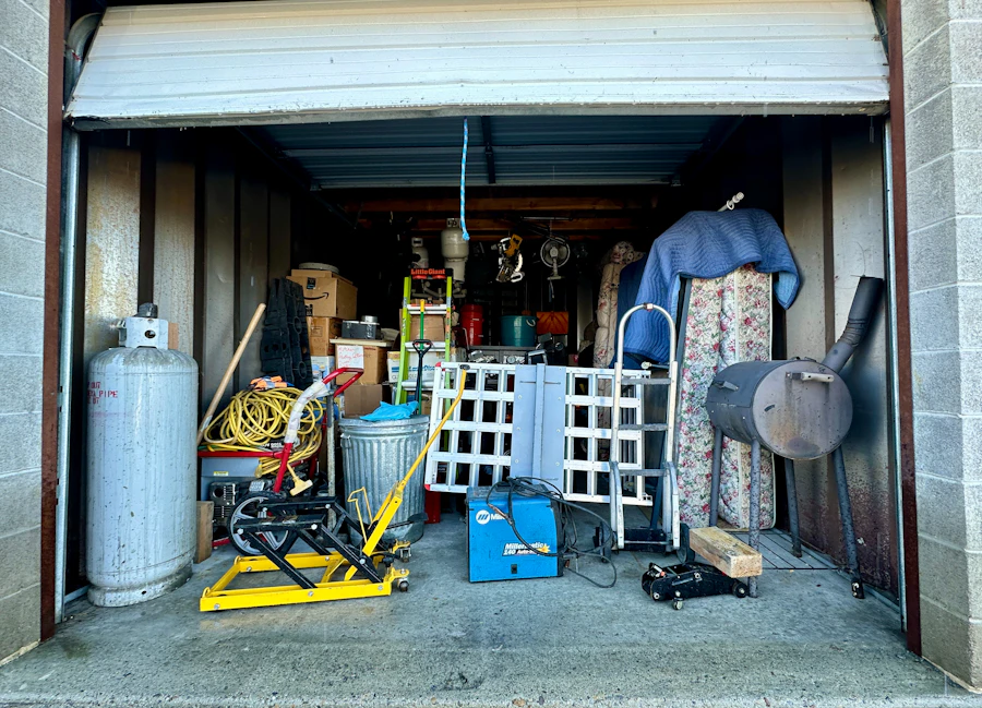 Packed self-storage unit with the roll-up door open, full of tools, boxes and old equipment