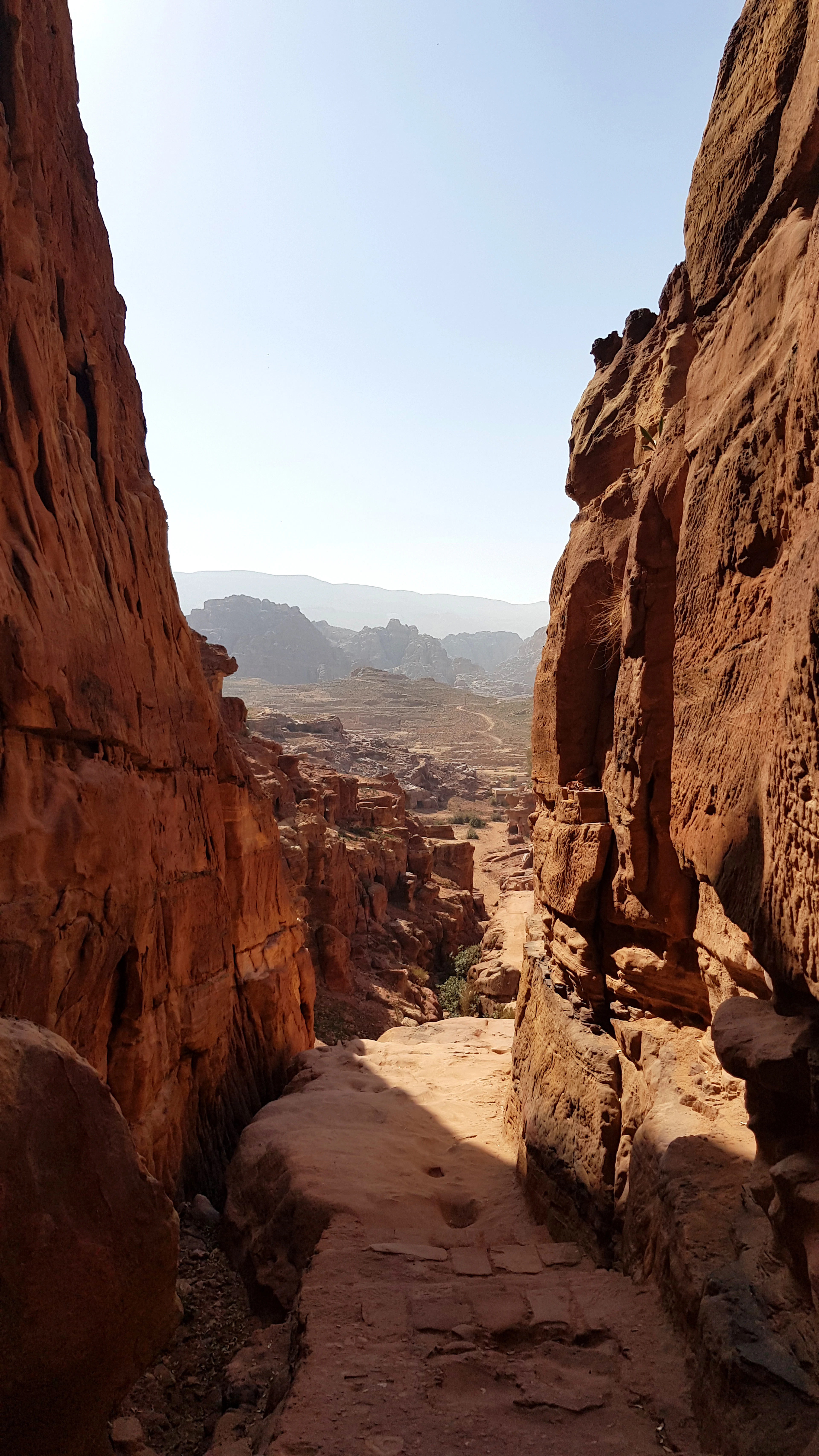 The rocky canyon trail up to Petra's Monastery, cut between red sandstone walls and opening onto the hazy rock basin beyond.