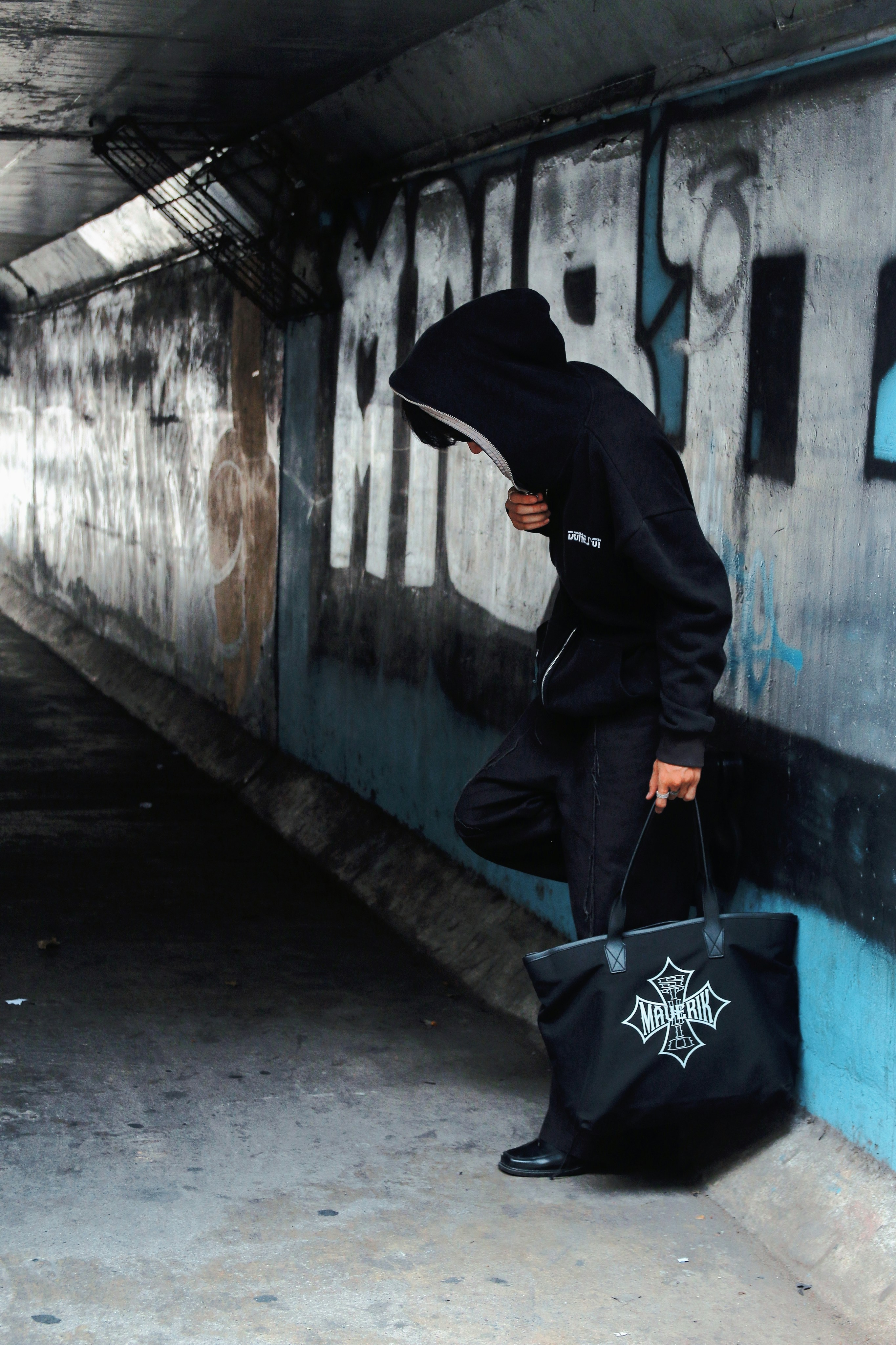 Person wearing oversized black hoodie in dark urban environment, low-key moody lighting, underground aesthetic
