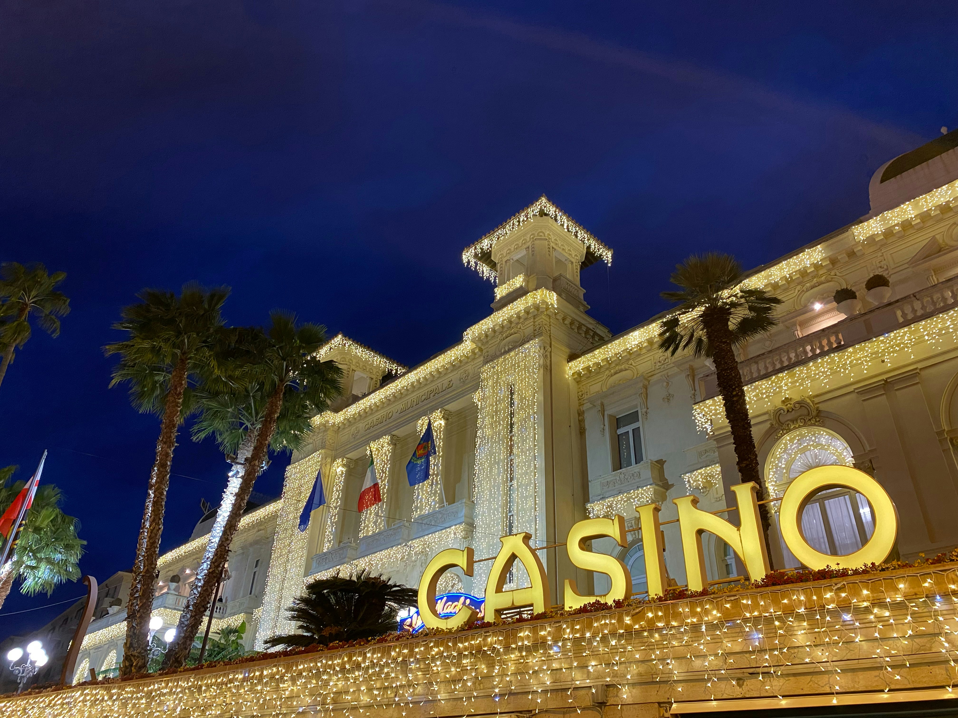 Grand casino resort interior with high ceilings, warm amber chandeliers, dark carpet with gold patterns, and a bustling gaming floor at night