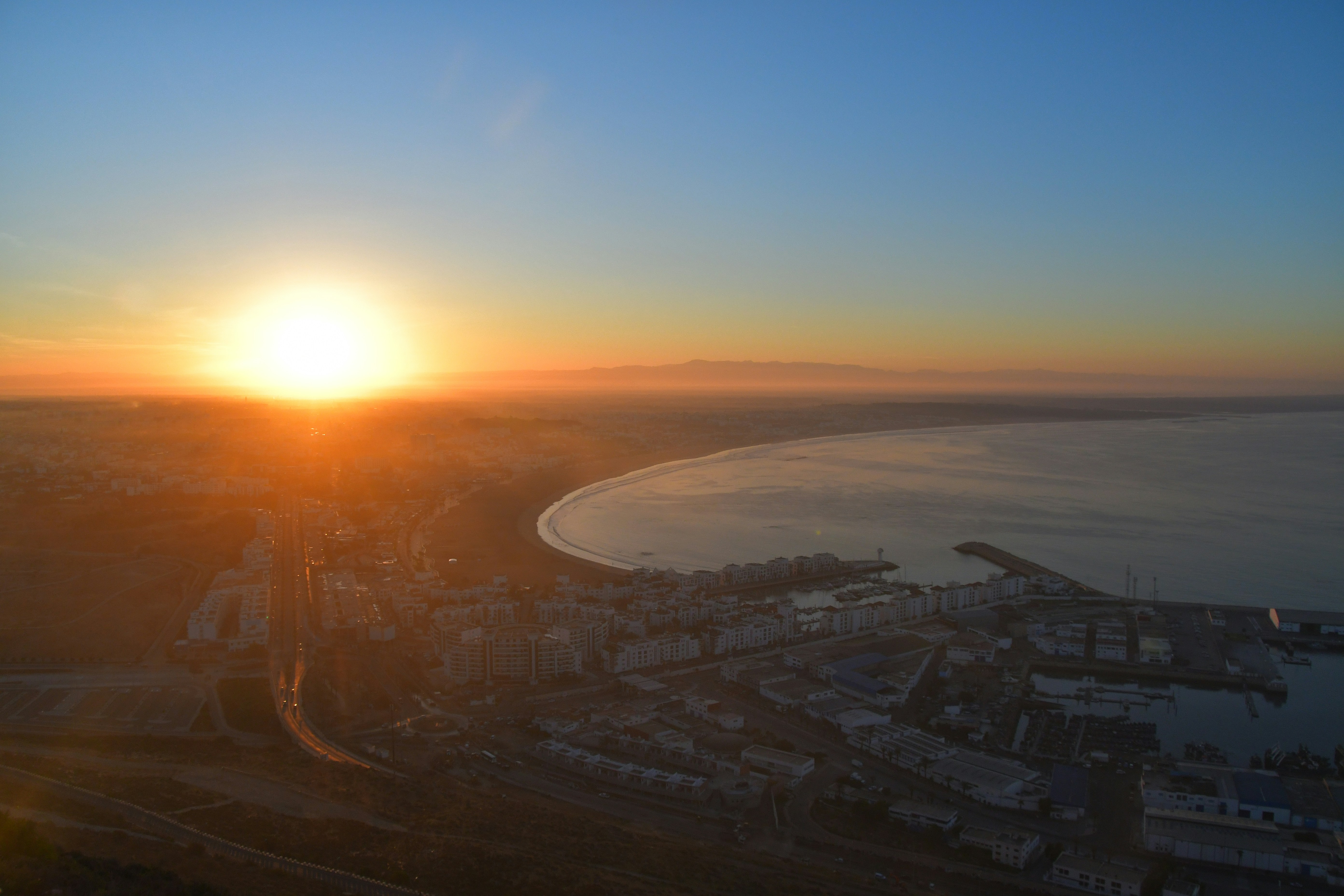 Aerial drone shot of coastal city at golden hour, warm amber and orange tones, long shadows, bright sky, dramatic landscape