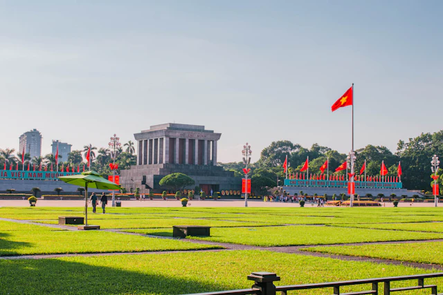 The Ho Chi Minh Mausoleum stands beyond a manicured lawn in Ba Dinh Square, flanked by a large Vietnamese flag.