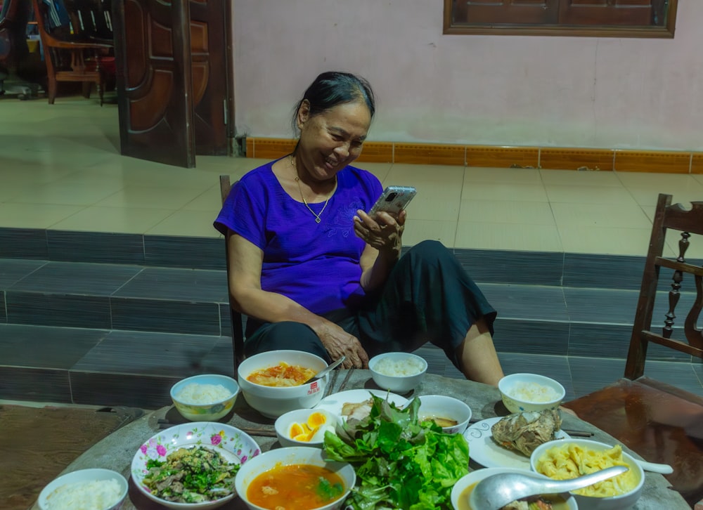 Una mujer sentada en una mesa llena de comida mirando su teléfono celular