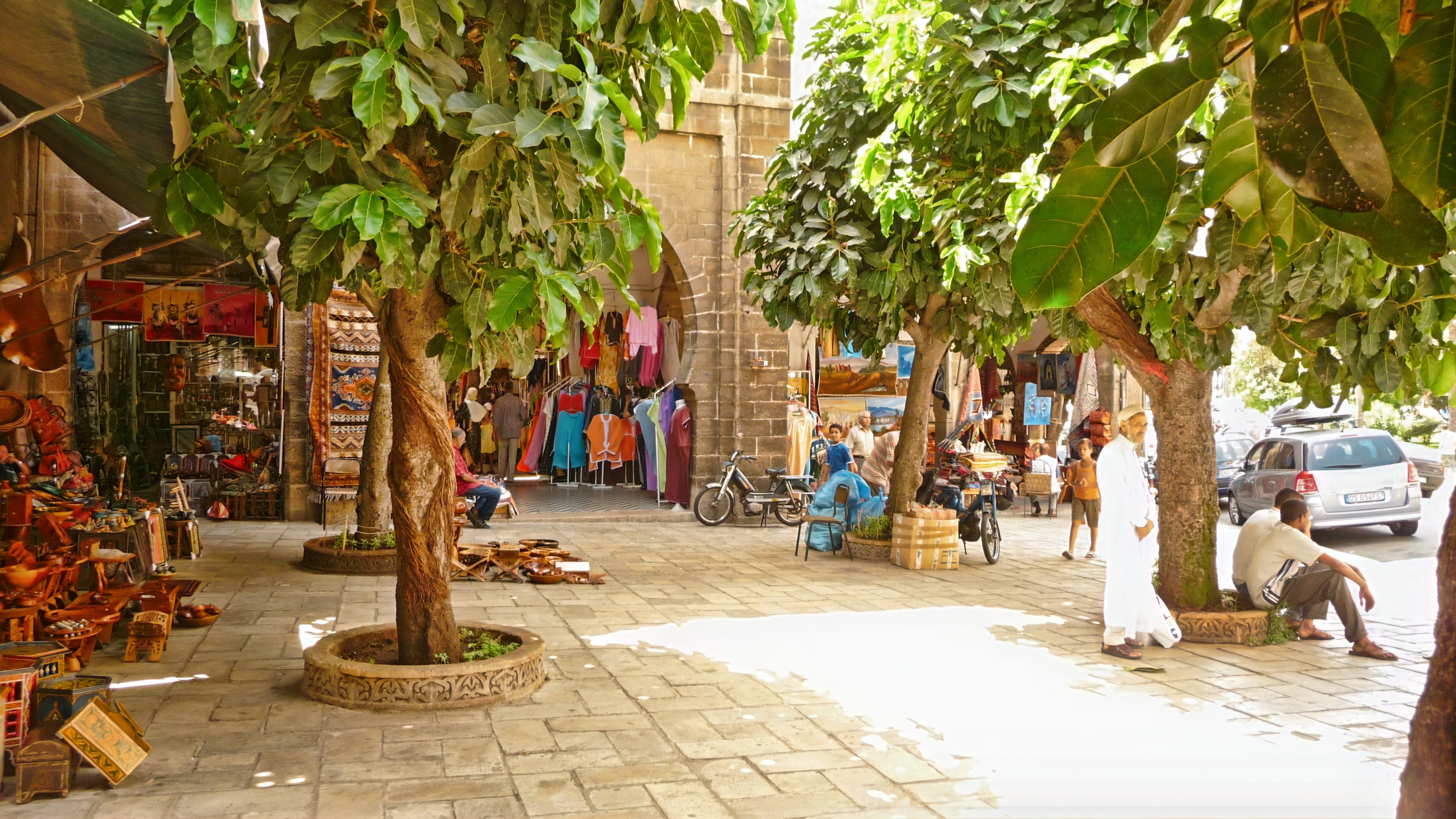 Shaded market alley in the Habous quarter, with shops, trees and shoppers under bright sun.