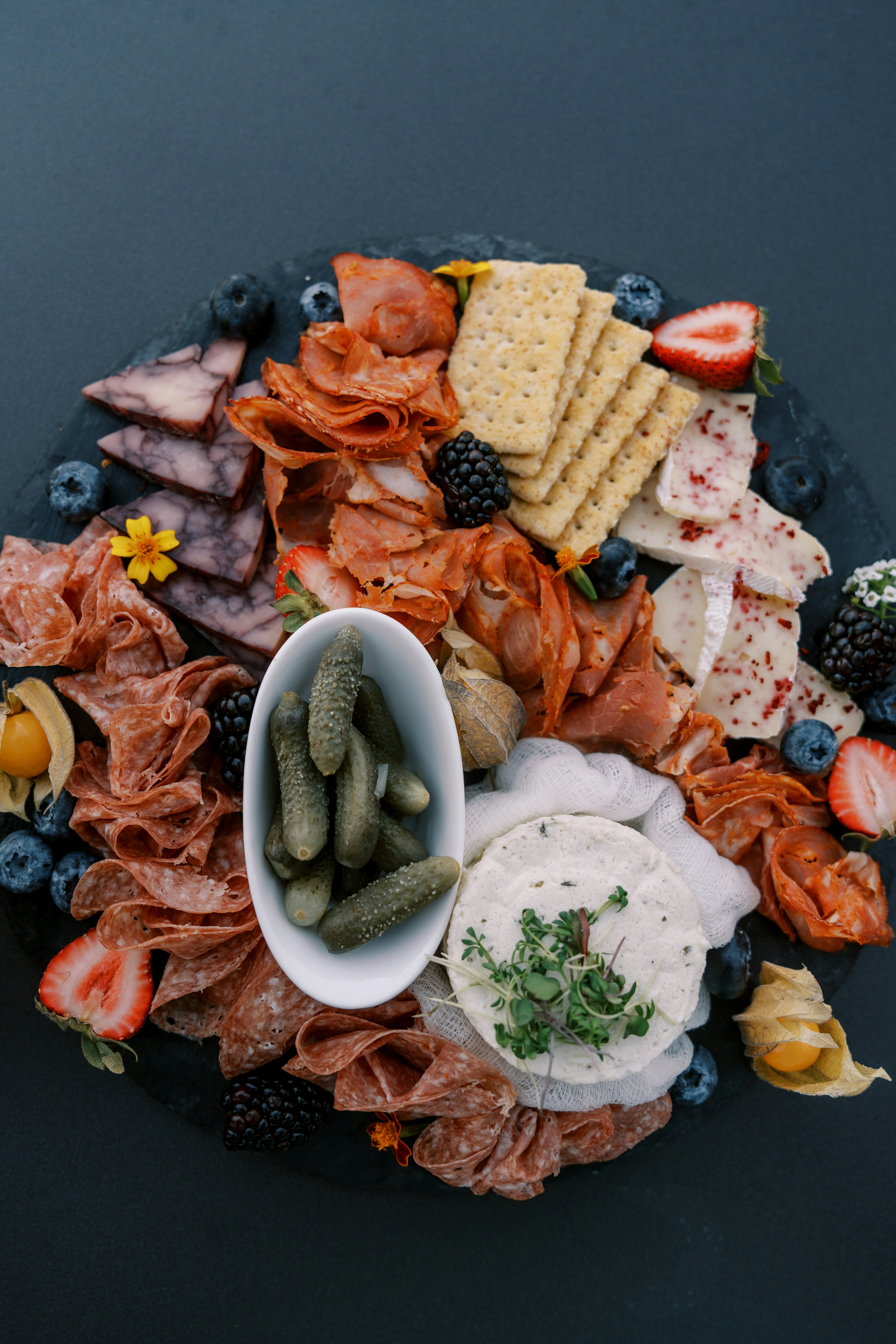 Artisan cheese and charcuterie board with dark accompaniments on slate surface in dim light