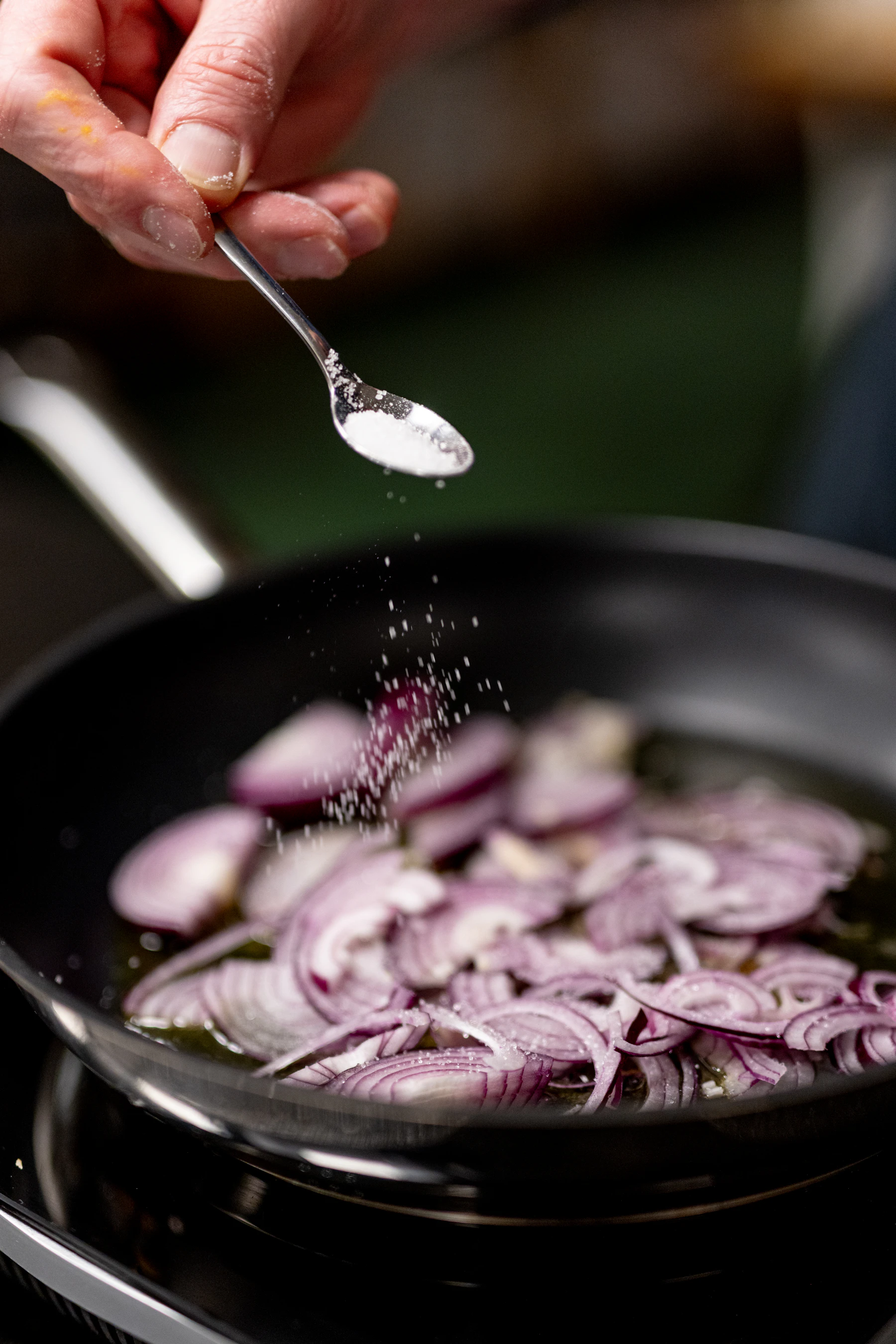 Sliced red onion softening in oil in a pan while salt falls from a spoon
