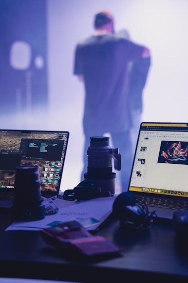 two laptops sitting on a desk with a man in the background