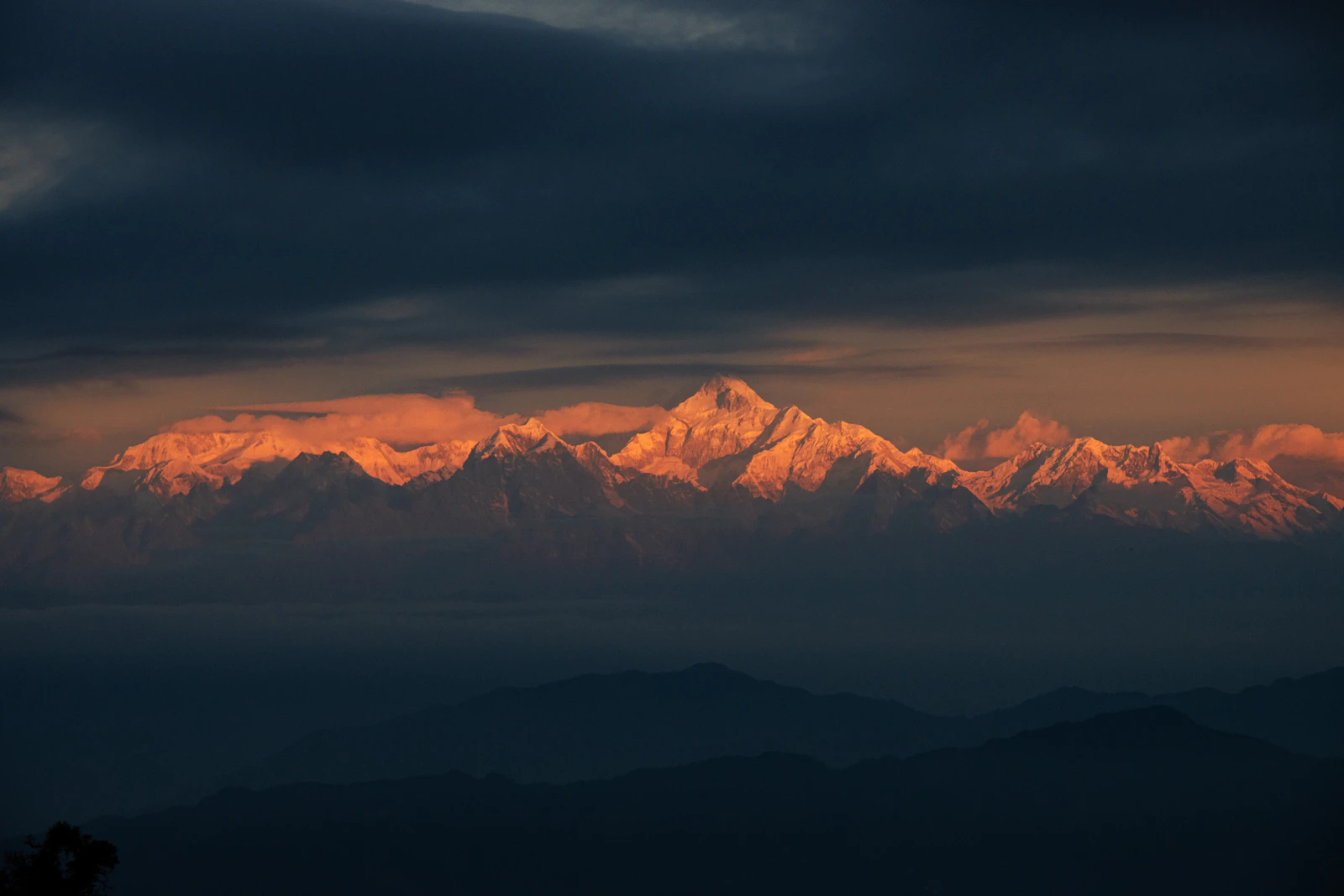 First light turning the Kanchenjunga range orange under a dark cloud bank, seen from Darjeeling above blue ridges