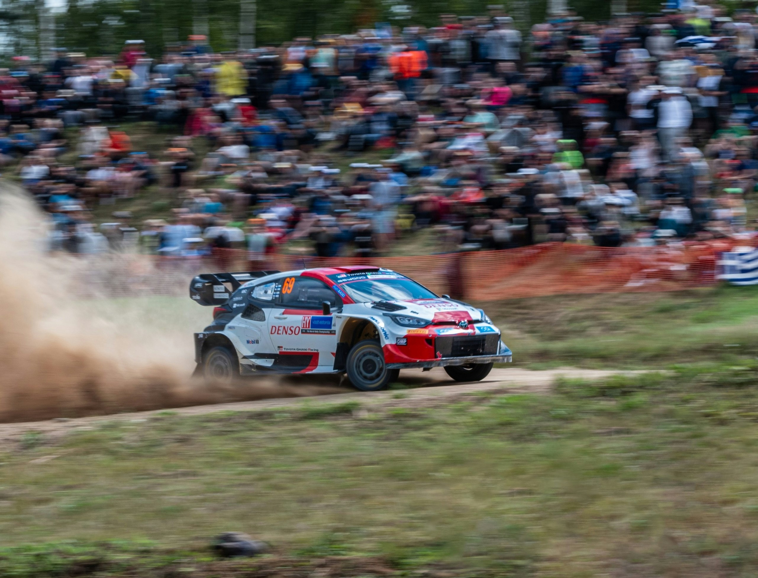 A white, with red and black Toyota Yaris WRC car going sideways on a gravel road with a wall of spectators behind watching from an embankment.