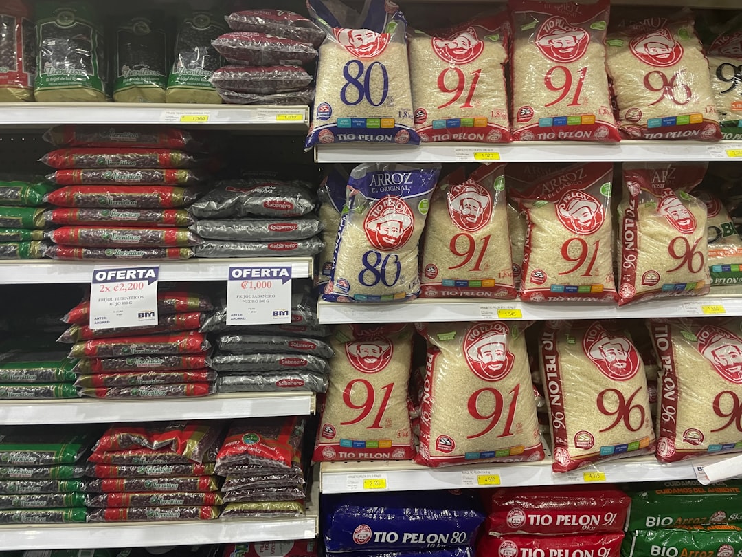 The image depicts a grocery store aisle stocked with bags of rice and beans, including brands like Tío Pelón. Sales offers are prominently displayed, highlighting the essentials for many households.