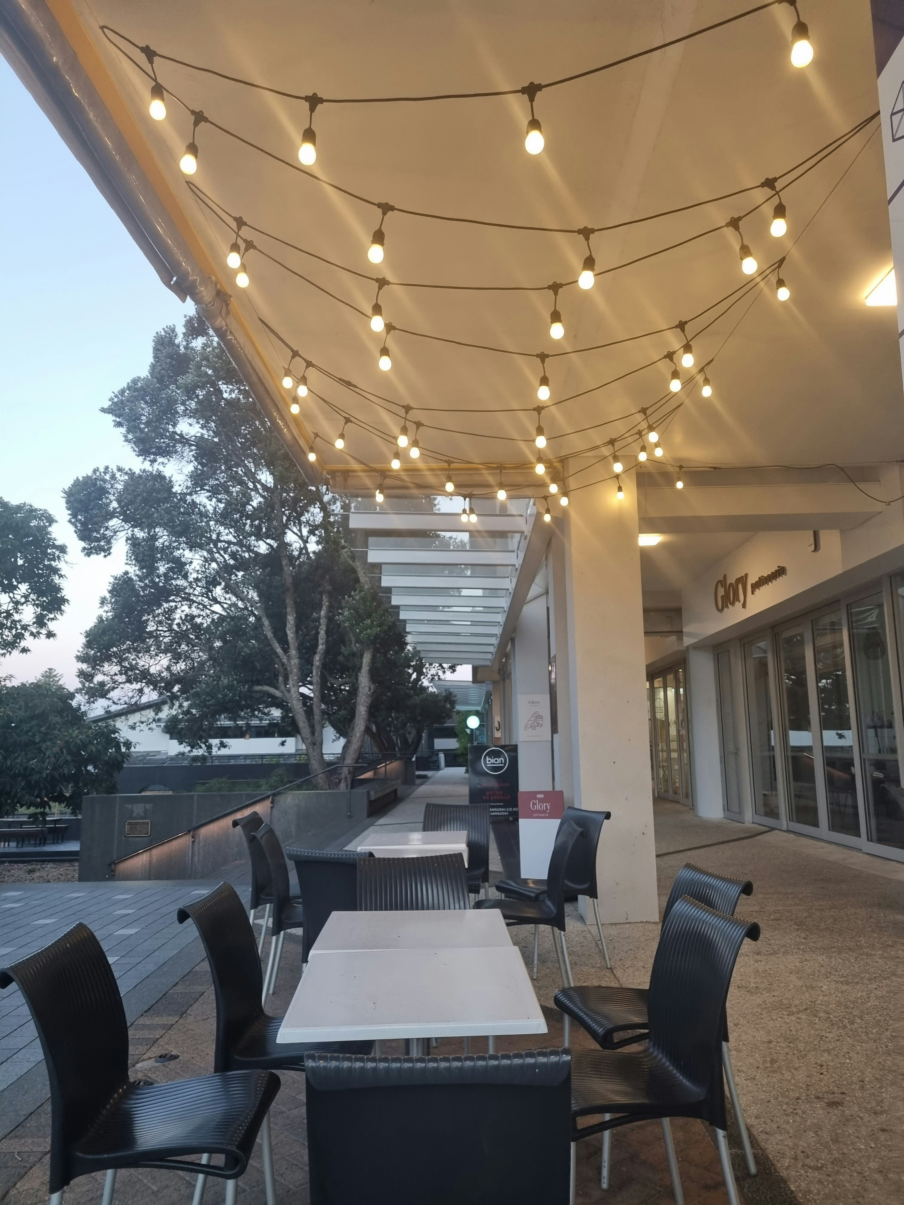 Cozy outdoor cafe seating area with string lights, potted plants, and people enjoying coffee in the evening