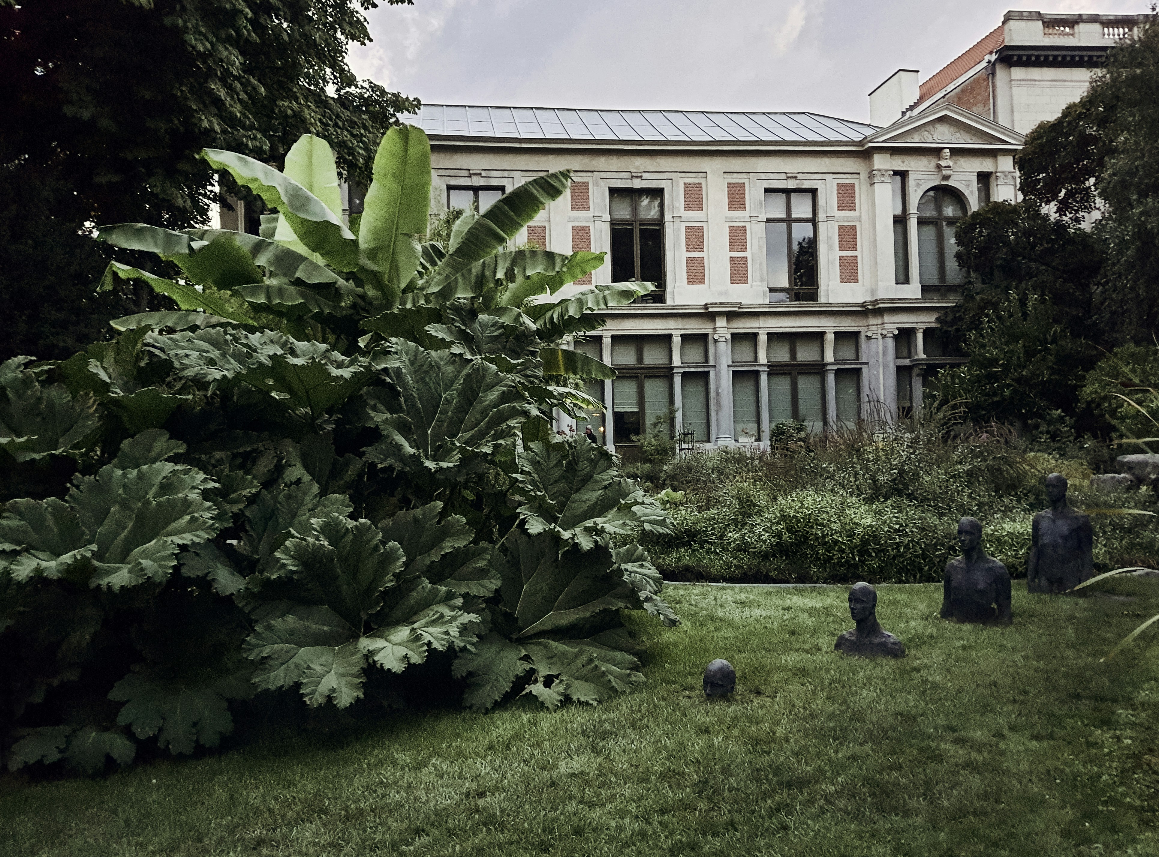 a large building sitting next to a lush green park, Statues of People in the Grass at Botanical Garden in Antwerp