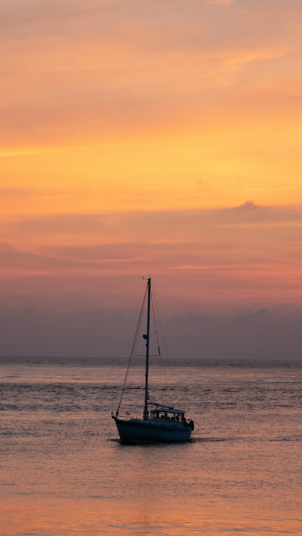 The image depicts a sailboat gently floating on the water under a beautiful sunset sky. The boat is positioned towards the right side of the frame, with its mast reaching up into the sky and its sails unfurled. The colors in the sky are vibrant shades of orange and yellow, creating a warm glow that contrasts with the cool blue of the ocean below. The horizon line is clearly visible in the background, adding depth to the scene.
