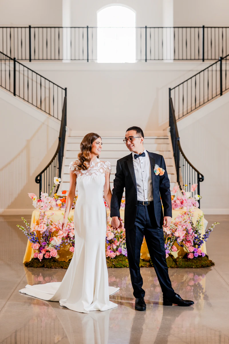 Pareja de novios tomada de la mano frente a una escalera decorada con flores