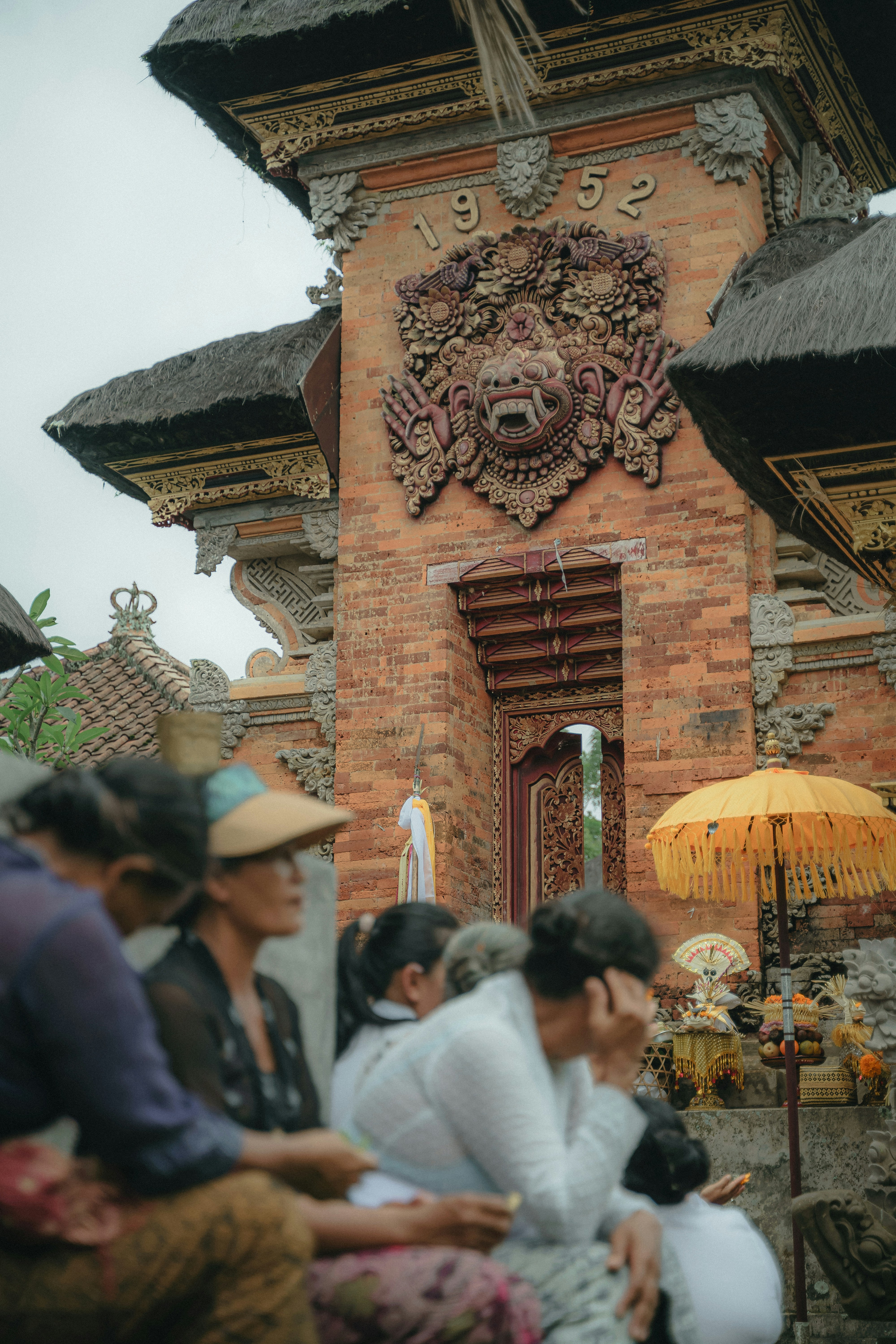 ocal Balinese women in traditional attire gather near the ornately carved entrance gate of a temple dated 1952 during a cultural ceremony — part of a rich Bali heritage tour experience.