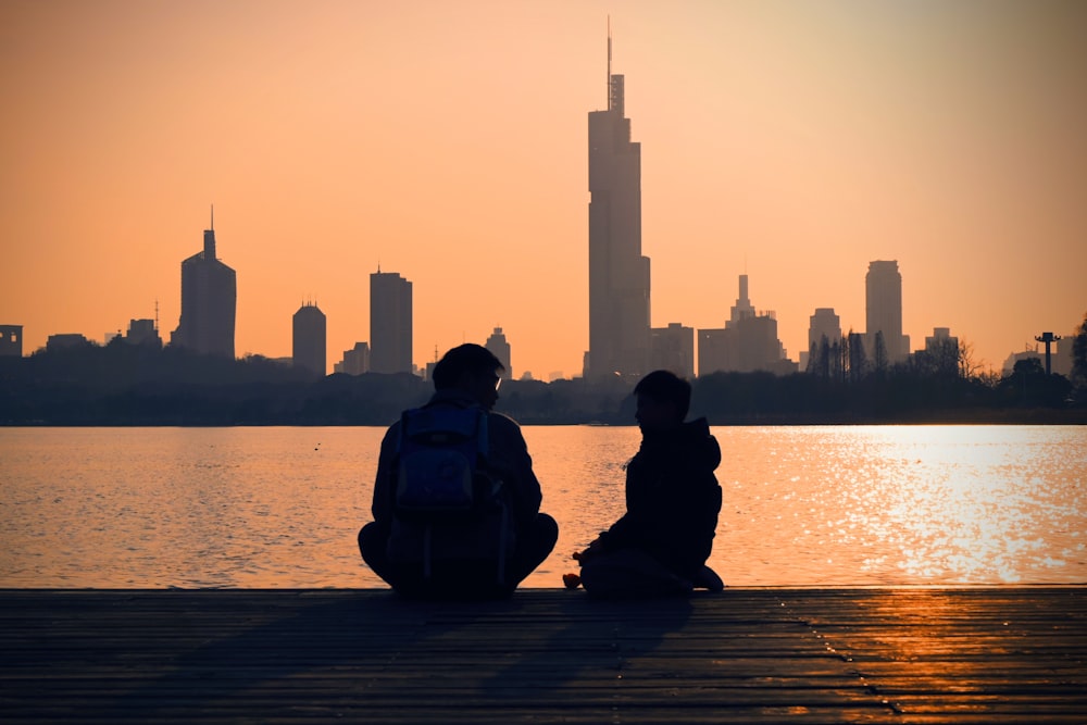 two people sitting on a dock in front of a body of water