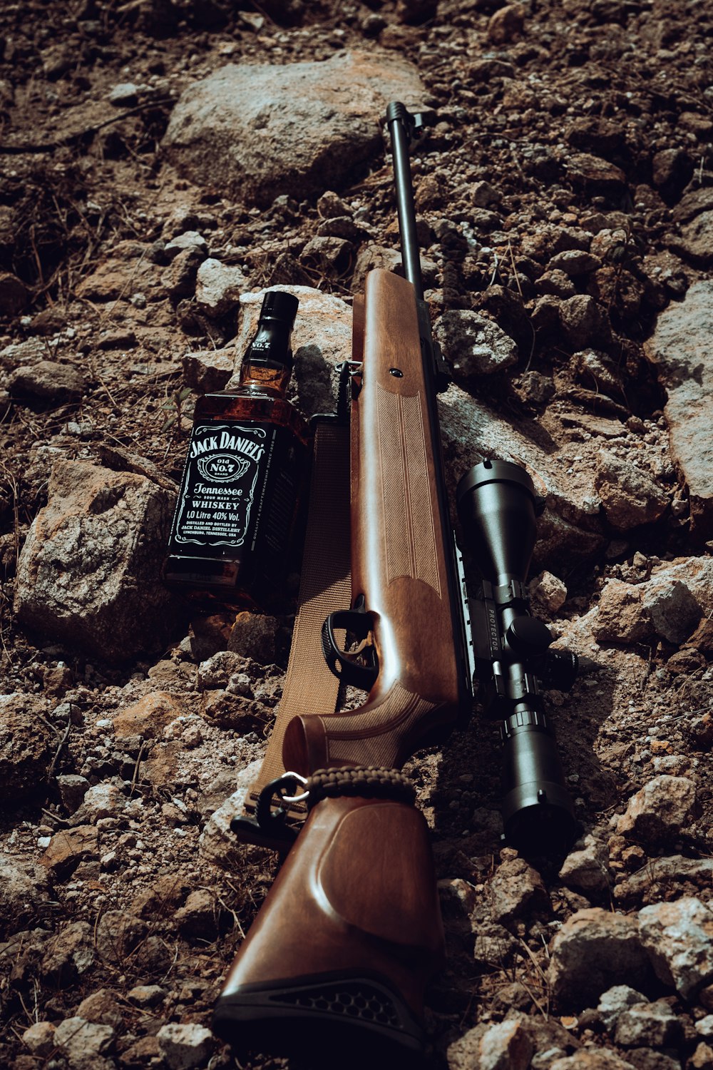 a rifle and a bottle of whiskey on a rocky ground