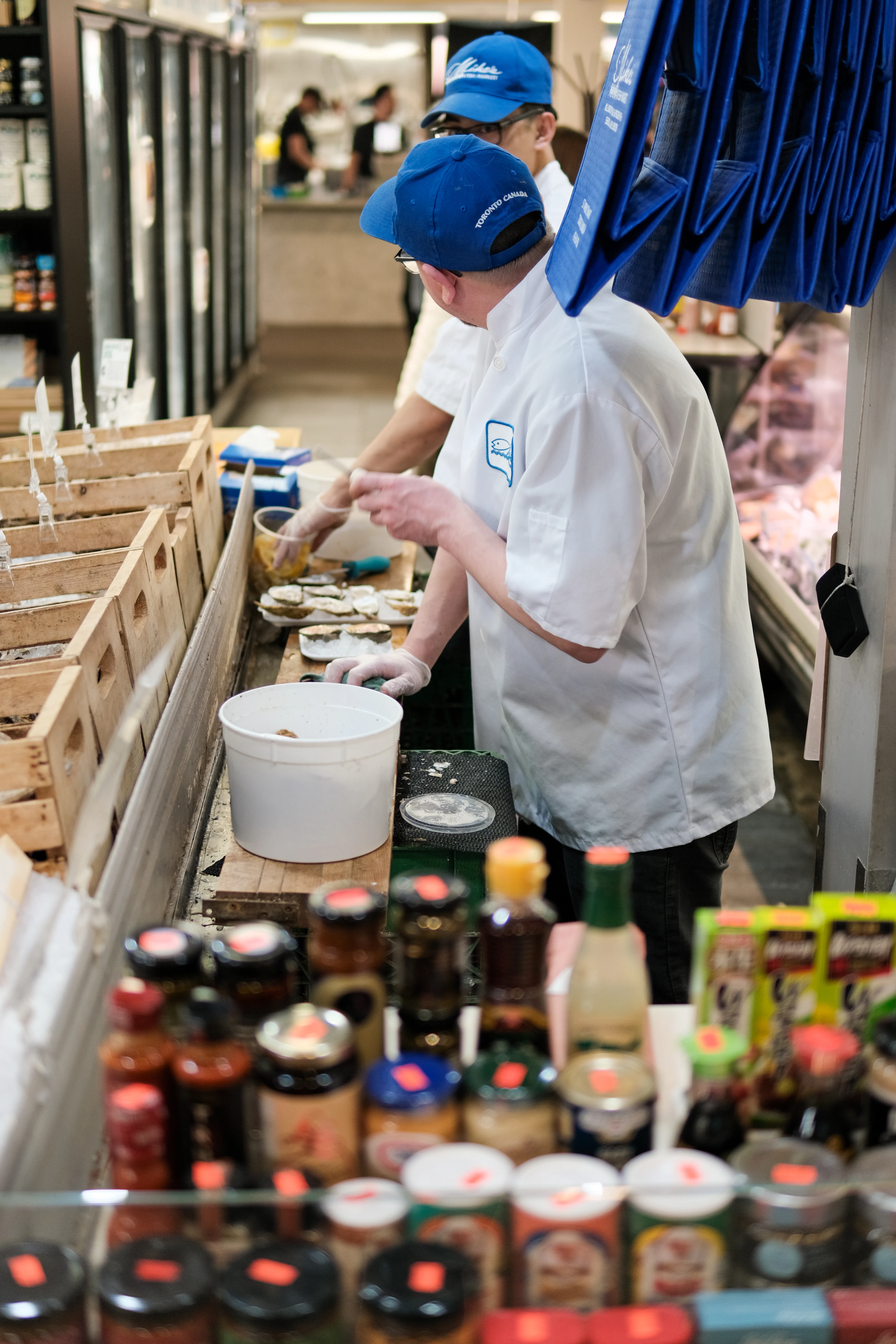 Fishmongers shucking oysters behind the counter at St Lawrence Market in Toronto, jars of preserves and hot sauces stacked along the front of the stall