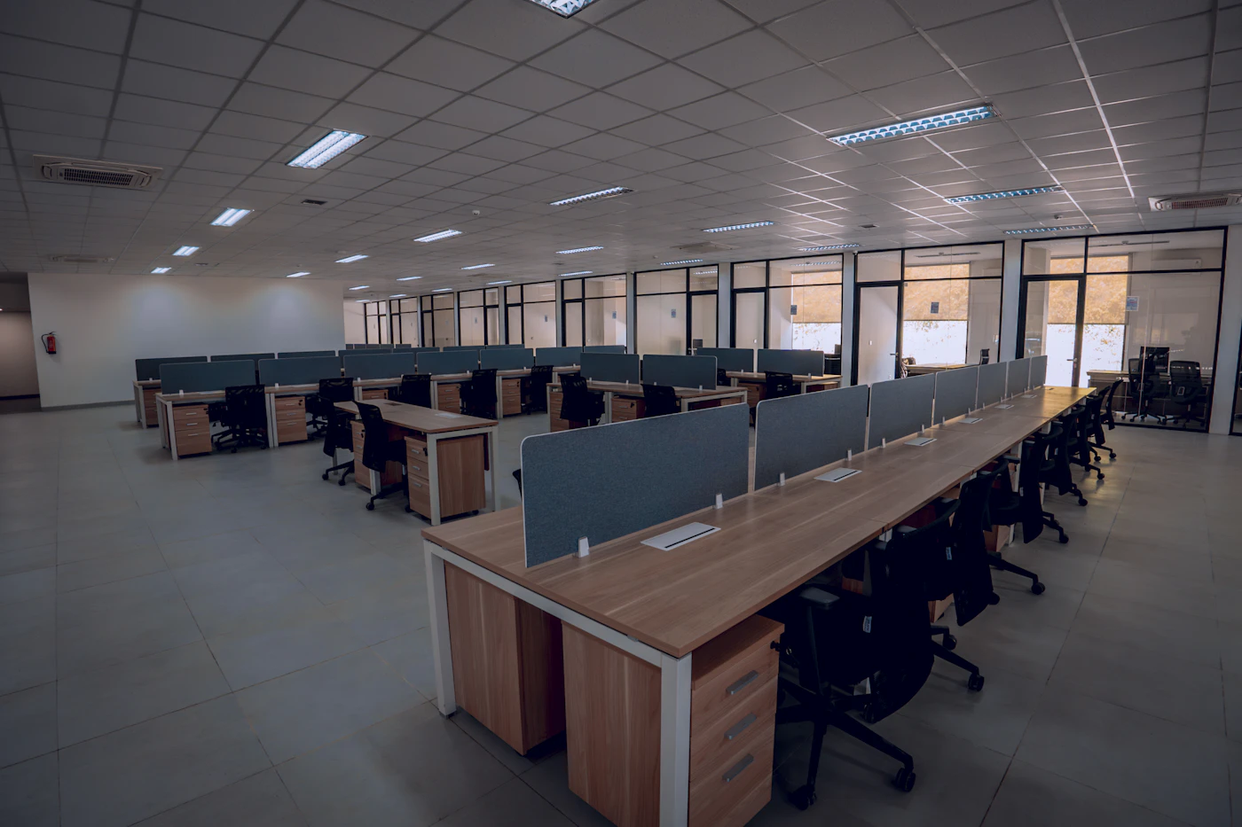An empty open-plan office of identical desks and chairs waiting under grey light, the workplace a return mandate sends people back to
