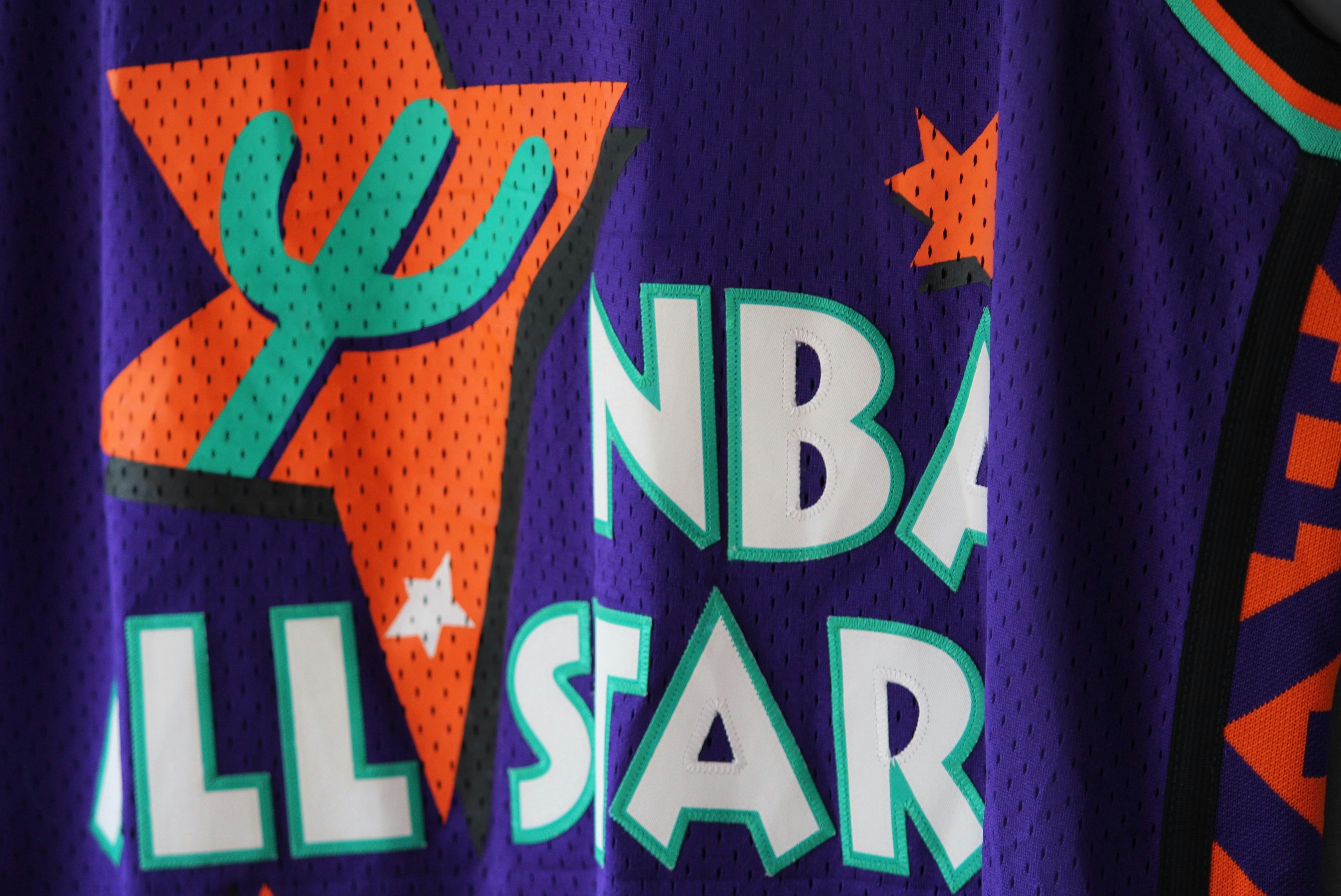 Close up image of a blue/purple NBA Adidas Soul Swingman 1994 East all star Penny Hardaway basketball jersey with teal and orange trims. Photo by Abhay Siby Mathew. Travel back to the 1995 NBA All-Star Game in Phoenix, where the jerseys rocked shades of purple, orange, bright teal, and black. These jerseys weren’t just about style; they were a tribute to the city’s vibe. Inspired by the Suns’ colors-purple, black, and orange-the designs also borrowed patterns from the Navajo culture, adding a cool twist to the game’s look. So, those 1995 jerseys weren’t just uniforms; they were a celebration of basketball’s connection to the vibrant tapestry of Phoenix’s culture and history. Photo by Abhay Siby Mathew.